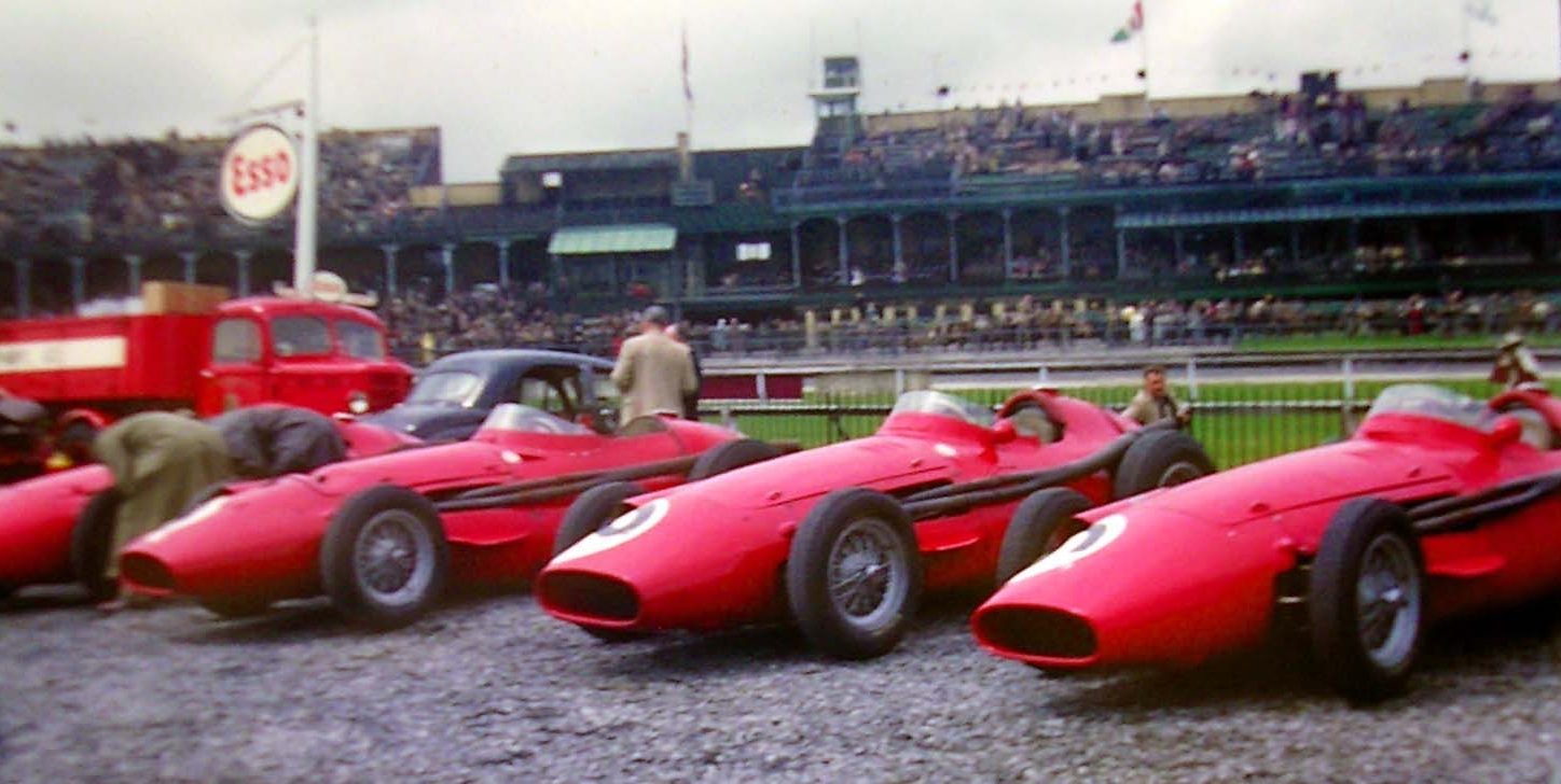 In colour: four red Maserati single-seaters parked side by side on gravel, their noses angled towards the camera, two men bent over the first two and two more standing among them; behind, a red lorry, a dark saloon car, an Esso sign and a crowded grandstand under a grey sky.