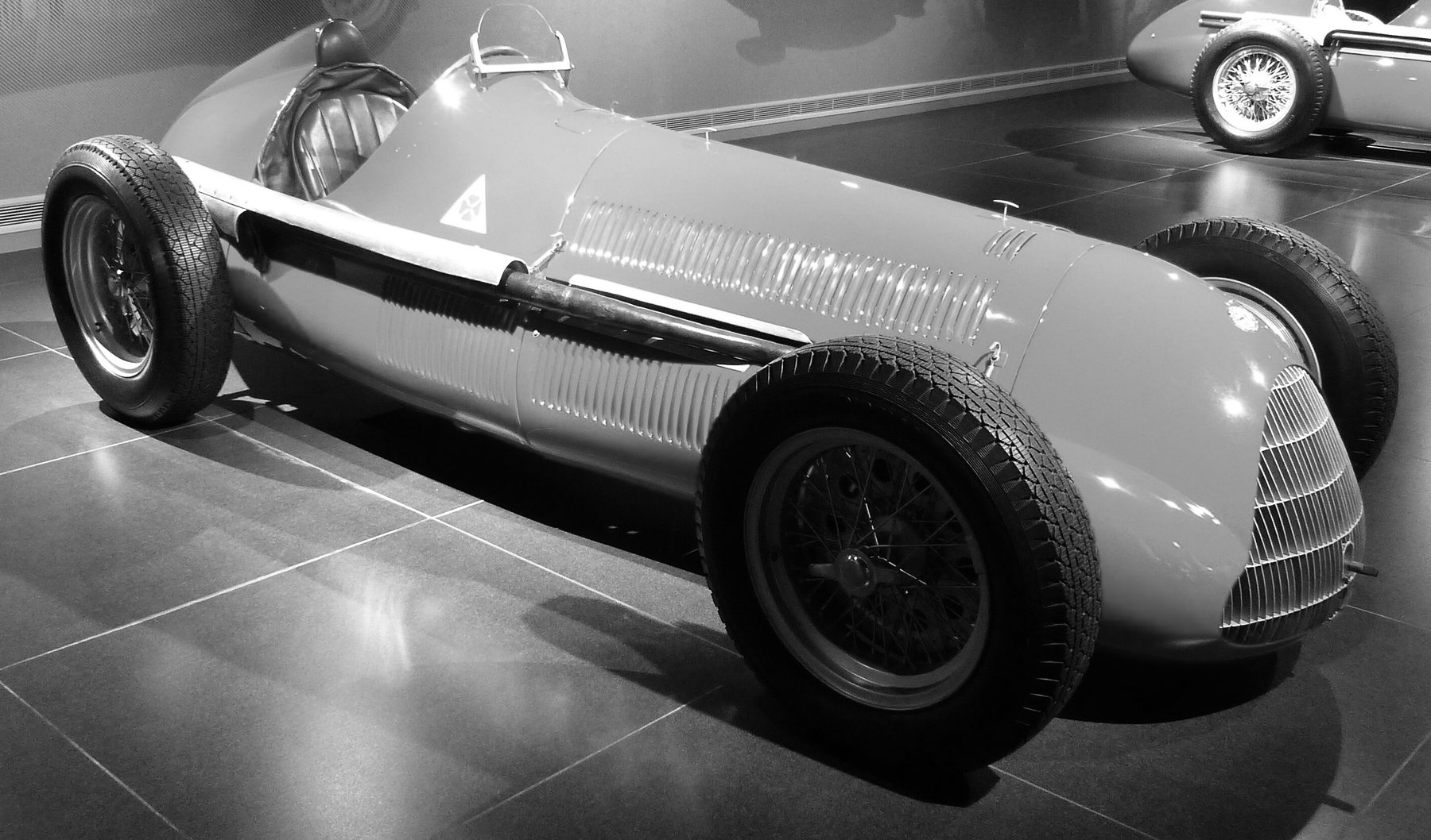 An Alfa Romeo single-seater in a museum, seen from the front three-quarters: a long rounded body with louvres along the bonnet, a grille of fine vertical bars, a small windscreen before an empty seat, an exhaust pipe along the flank and a four-leaf clover in a triangle on the cowl; part of a second single-seater stands behind it at the right.