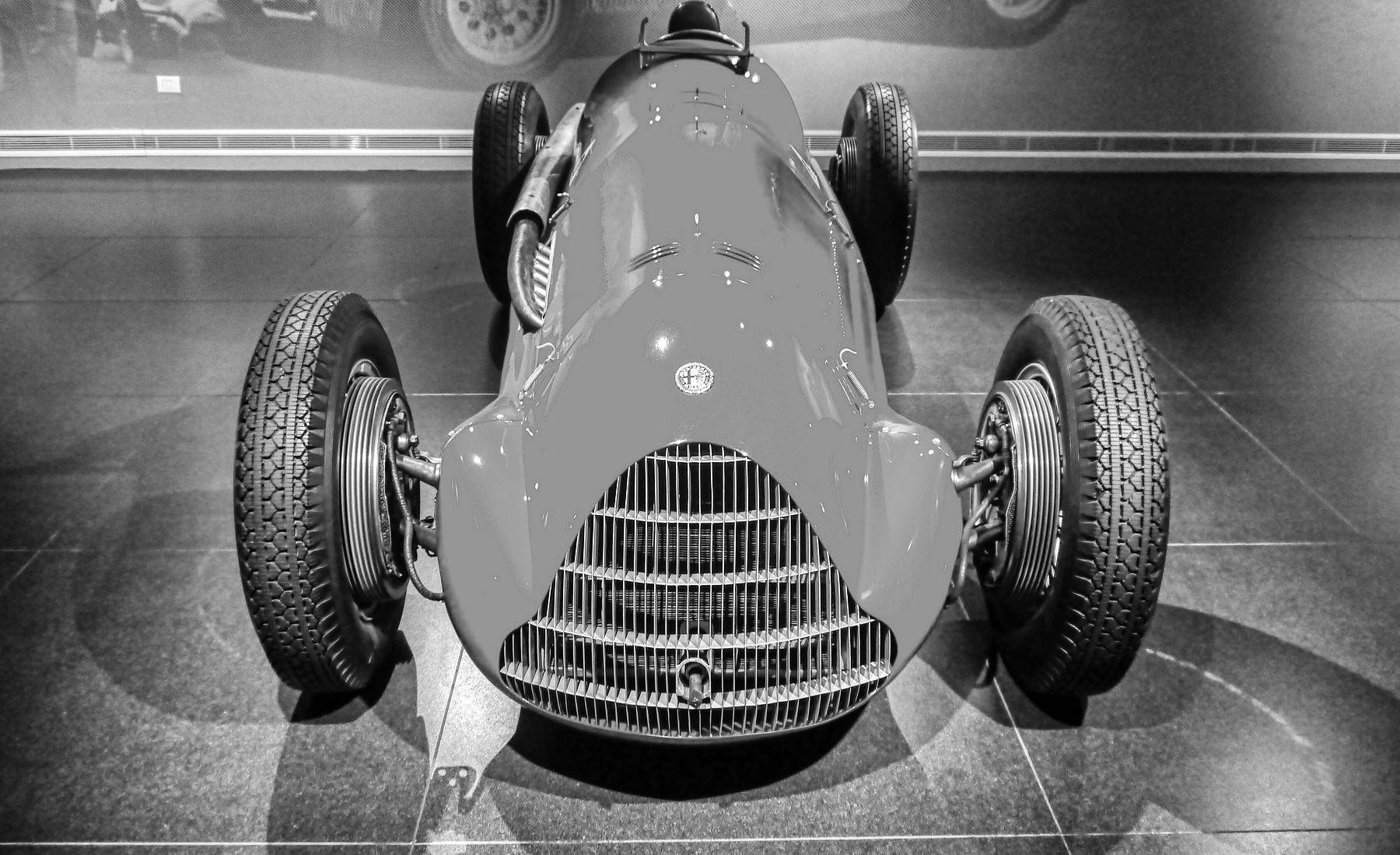 Head-on and close: an Alfa Romeo single-seater with a tall grille of vertical bars, a round badge on its long rounded bonnet, bare front brake drums and wire wheels, on a dark tiled floor; a wall of enlarged period photographs soft behind it.