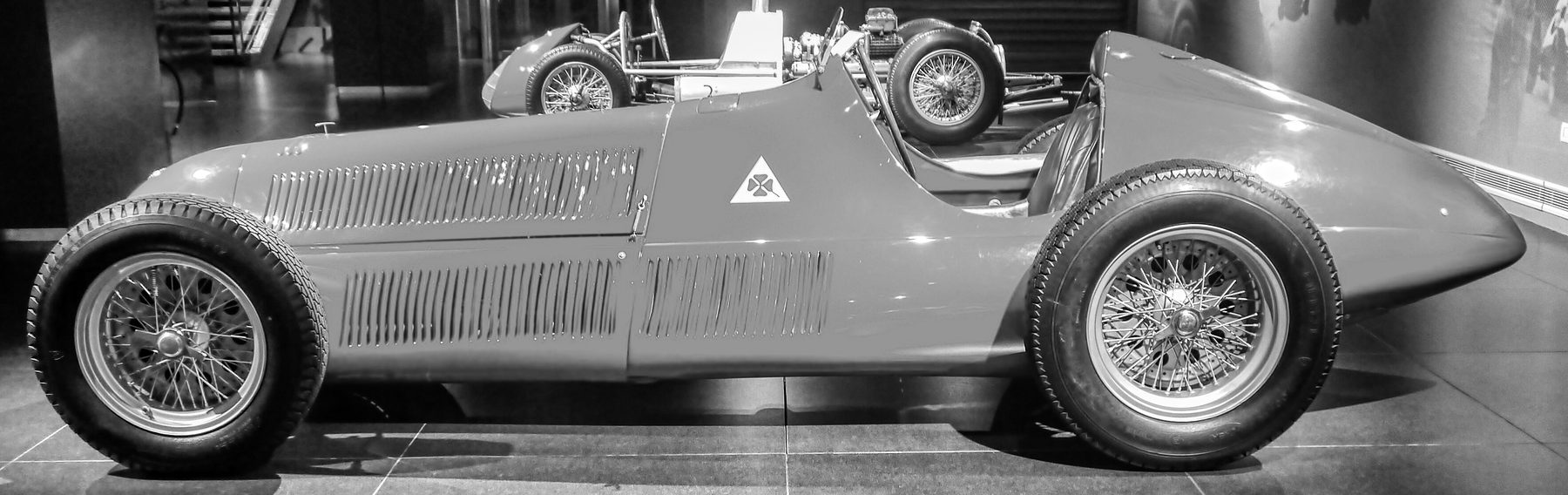 A single-seater in full profile, nose to the left, with rows of louvres along its bonnet, a four-leaf clover in a white triangle on the scuttle, a curved windscreen, a long tapering tail and wire wheels, on a dark reflecting floor; the front of another racing car and the frame of a bare chassis stand behind it.