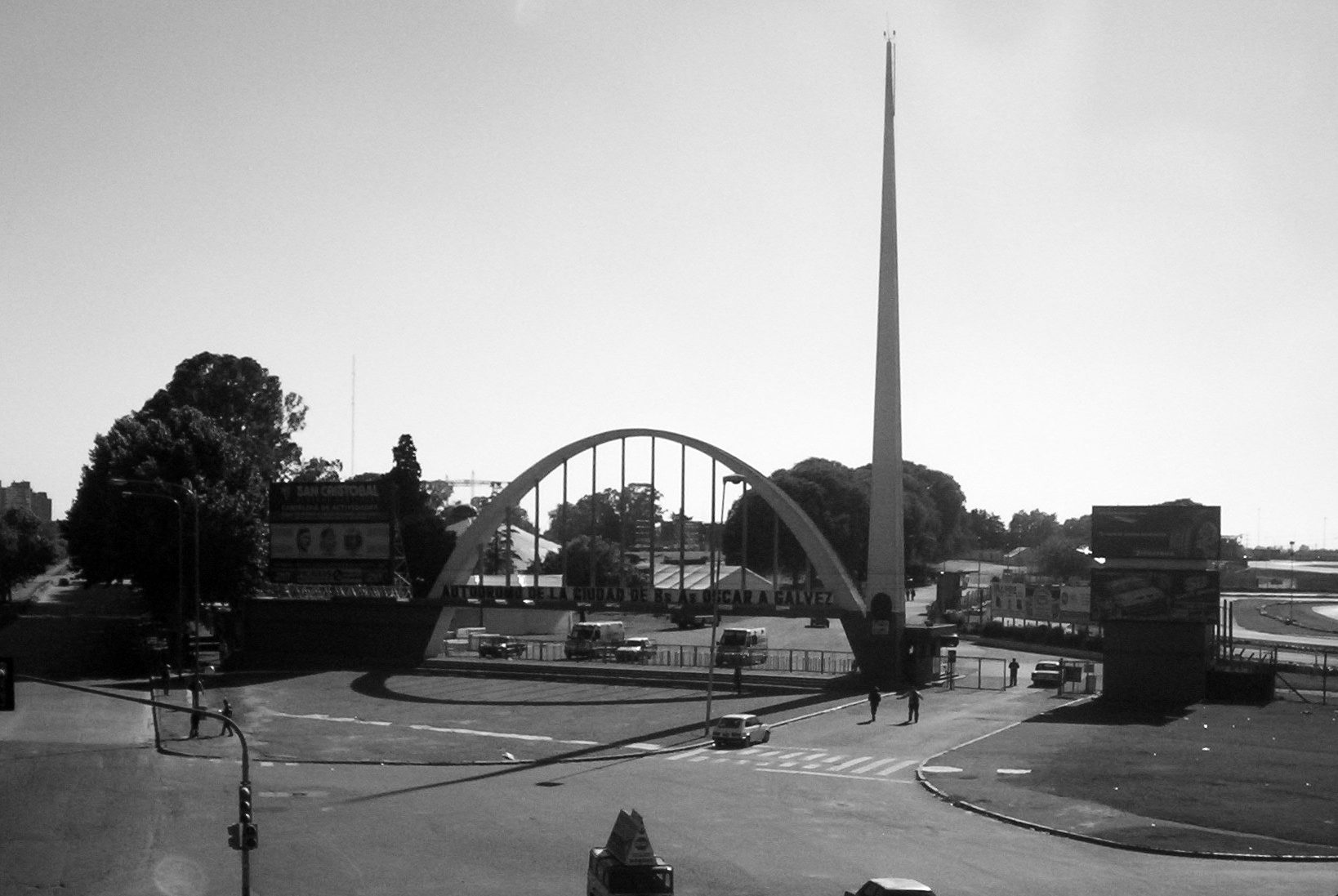 The entrance to a motor-racing circuit seen from a footbridge: a white arch over the gates and a tall, slender spire beside it, trees and a wide road junction in front.