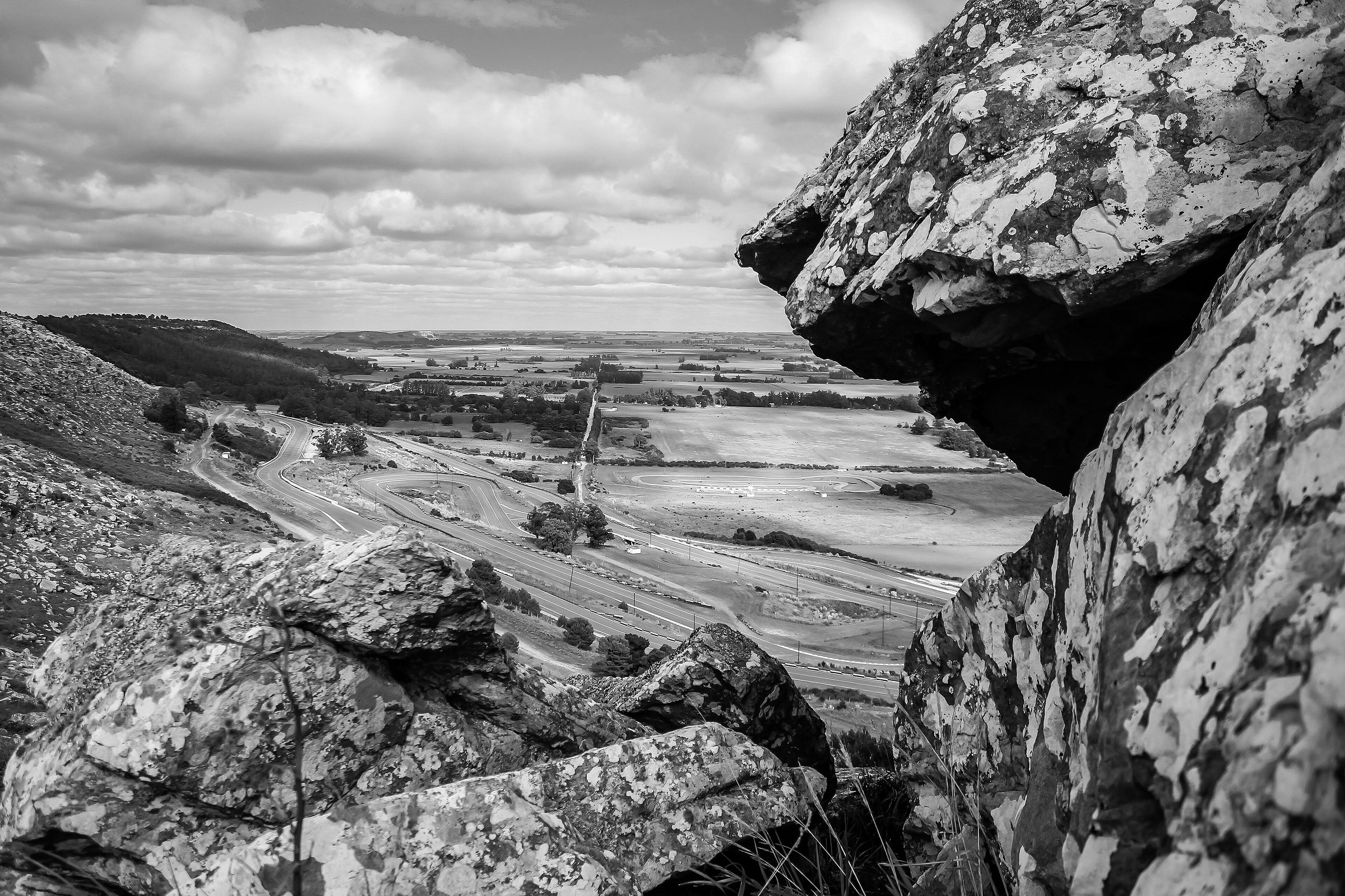 From among lichen-covered boulders high on a hillside, a view down to a winding motor-racing circuit at the foot of the slope and, beyond it, flat farmland of fields and tree lines stretching to the horizon under a big sky.
