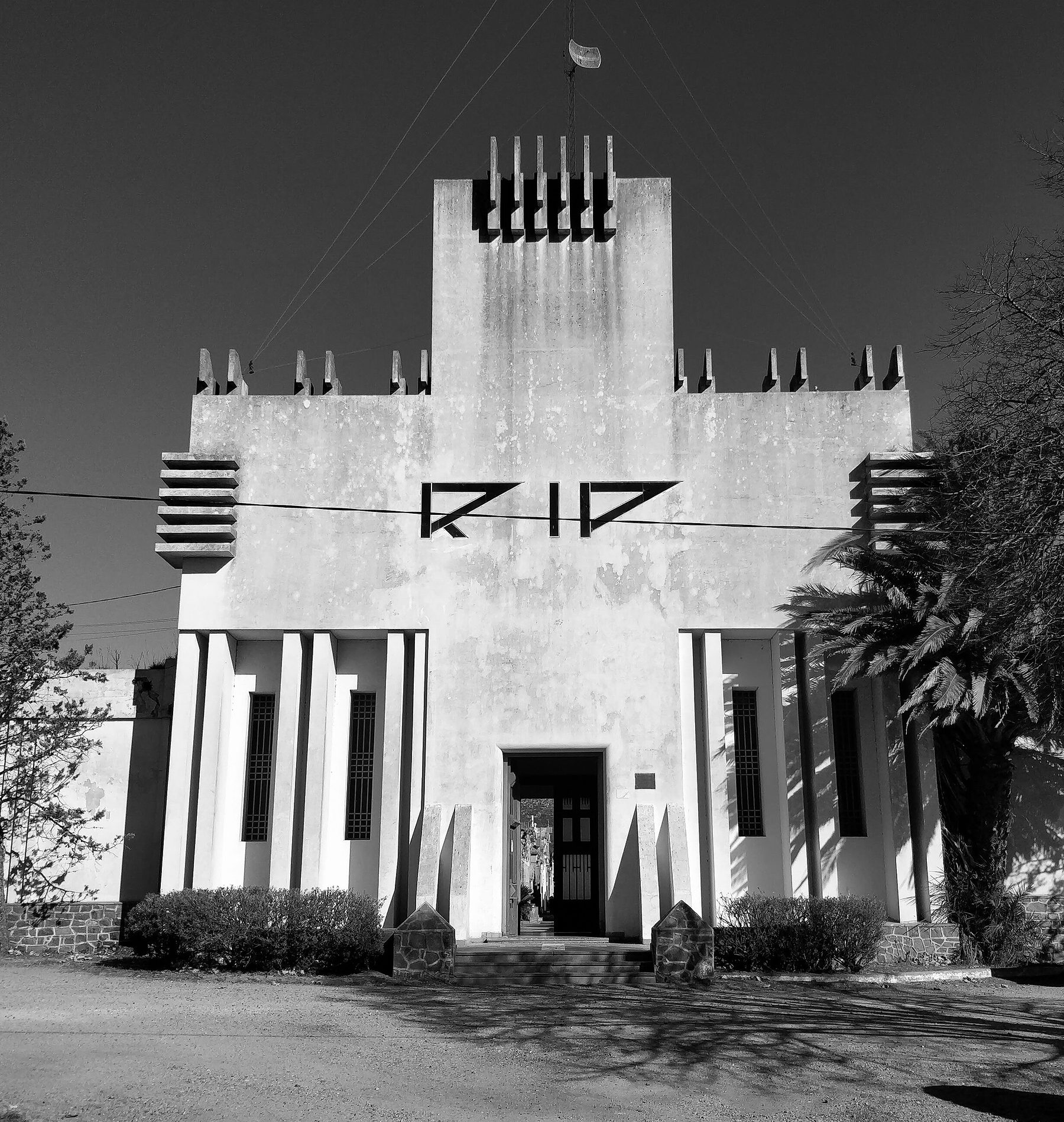 The white Art Deco gateway of a cemetery under a clear sky: a tall central tower crowned with comb-like crenellations, the letters RIP in angular capitals, vertical fins either side of a narrow doorway, crenellated walls, a palm and pines.