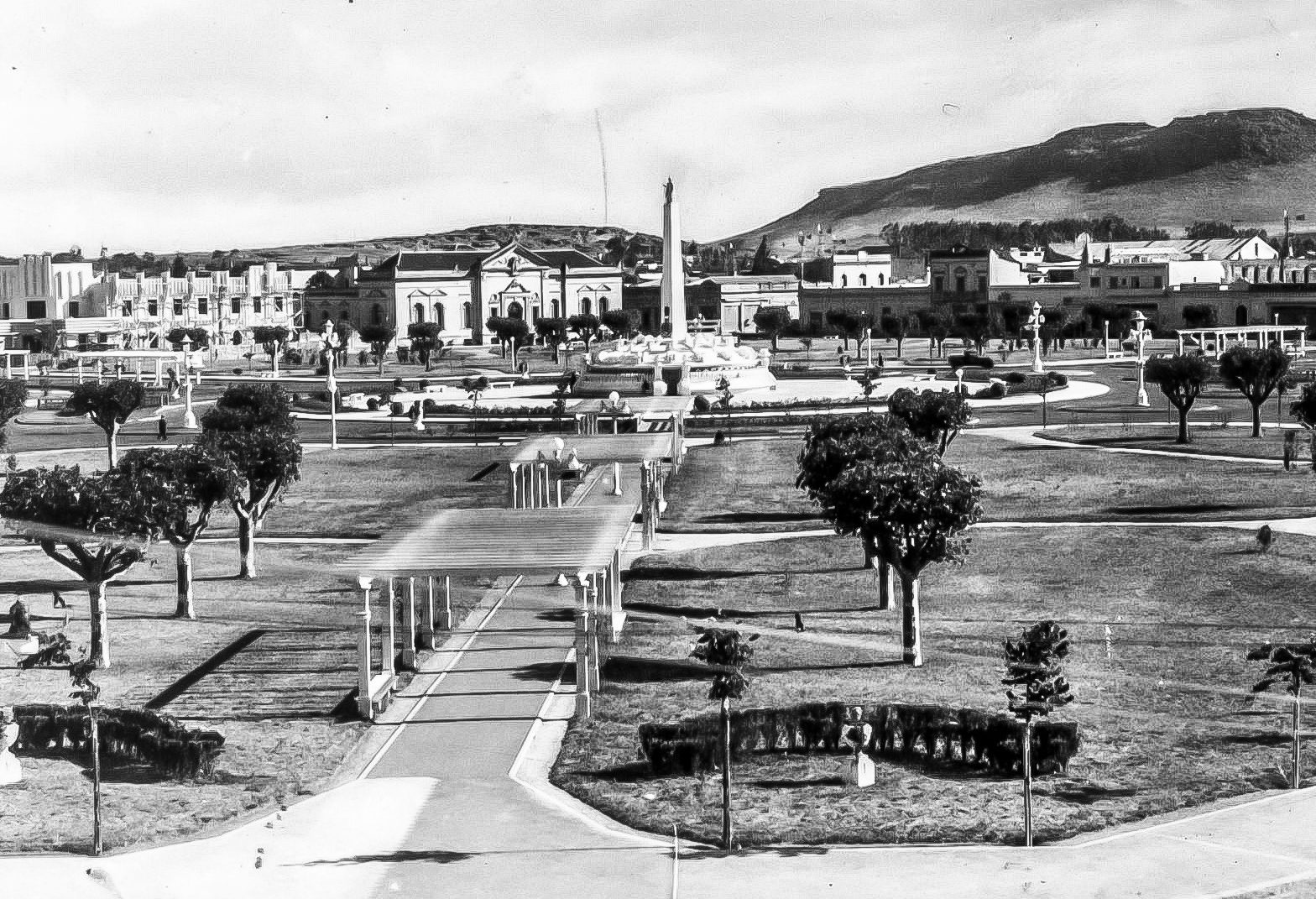An old postcard view over a newly laid-out square: young clipped trees, a pergola walk, lamp standards and an obelisk with a fountain at its centre, a row of low buildings beyond and a long bare ridge of hills behind the town.