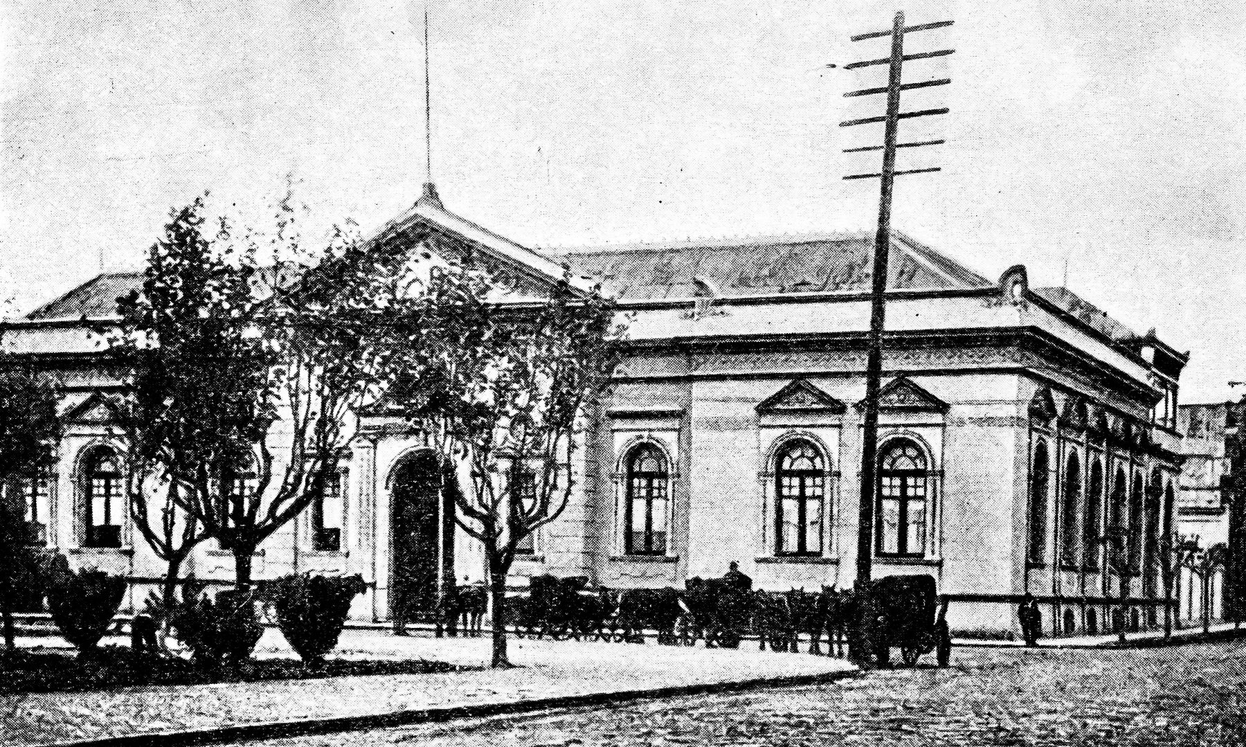 A book halftone of a single-storey public building on a street corner, with arched windows and a central pediment, young trees in front, a line of horse-drawn carriages waiting along the kerb and a tall telegraph pole.