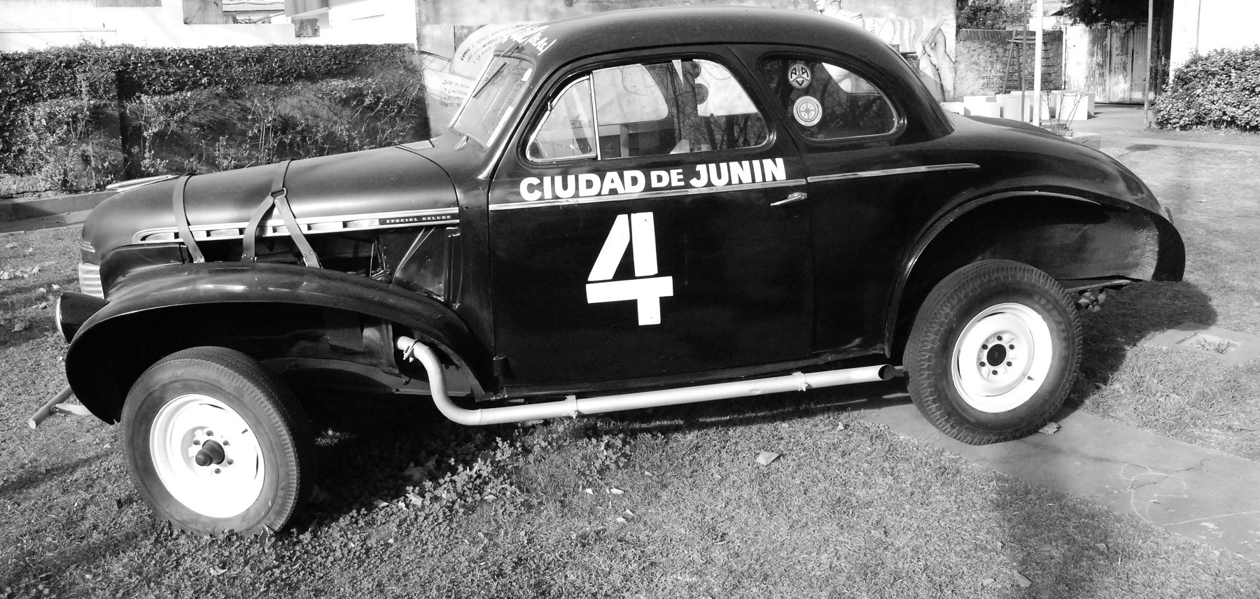 A late-1930s coupé prepared for road racing stands side-on on a lawn: dark paint, CIUDAD DE JUNIN and a big 4 on the door, leather straps across the bonnet, a straight exhaust pipe along the sill, and pale wheels.