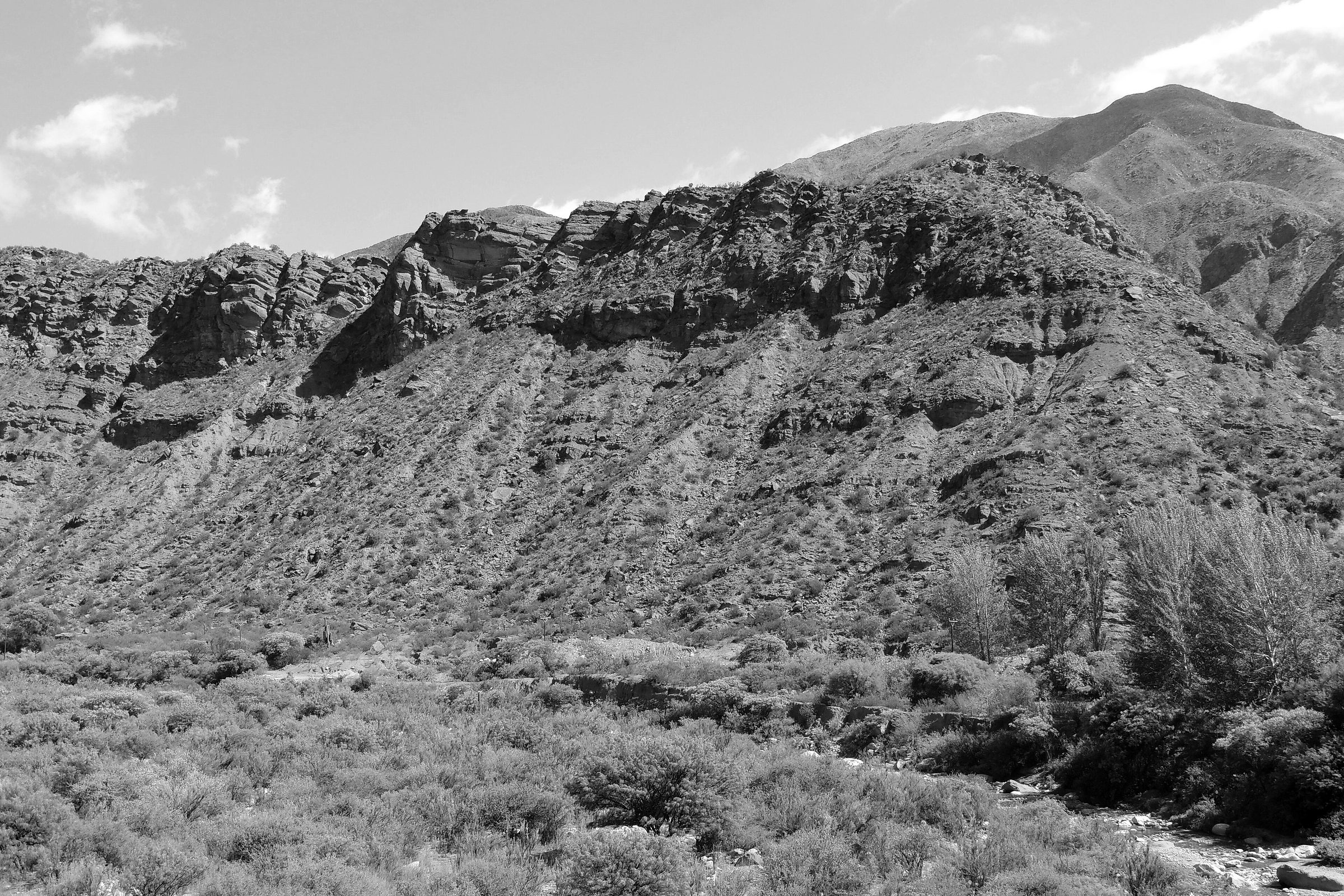 A great slope of rough sandstone rises above a scrubby river bed, bare ridges of rock along its crest and higher mountains behind, under a clear sky with a few clouds.