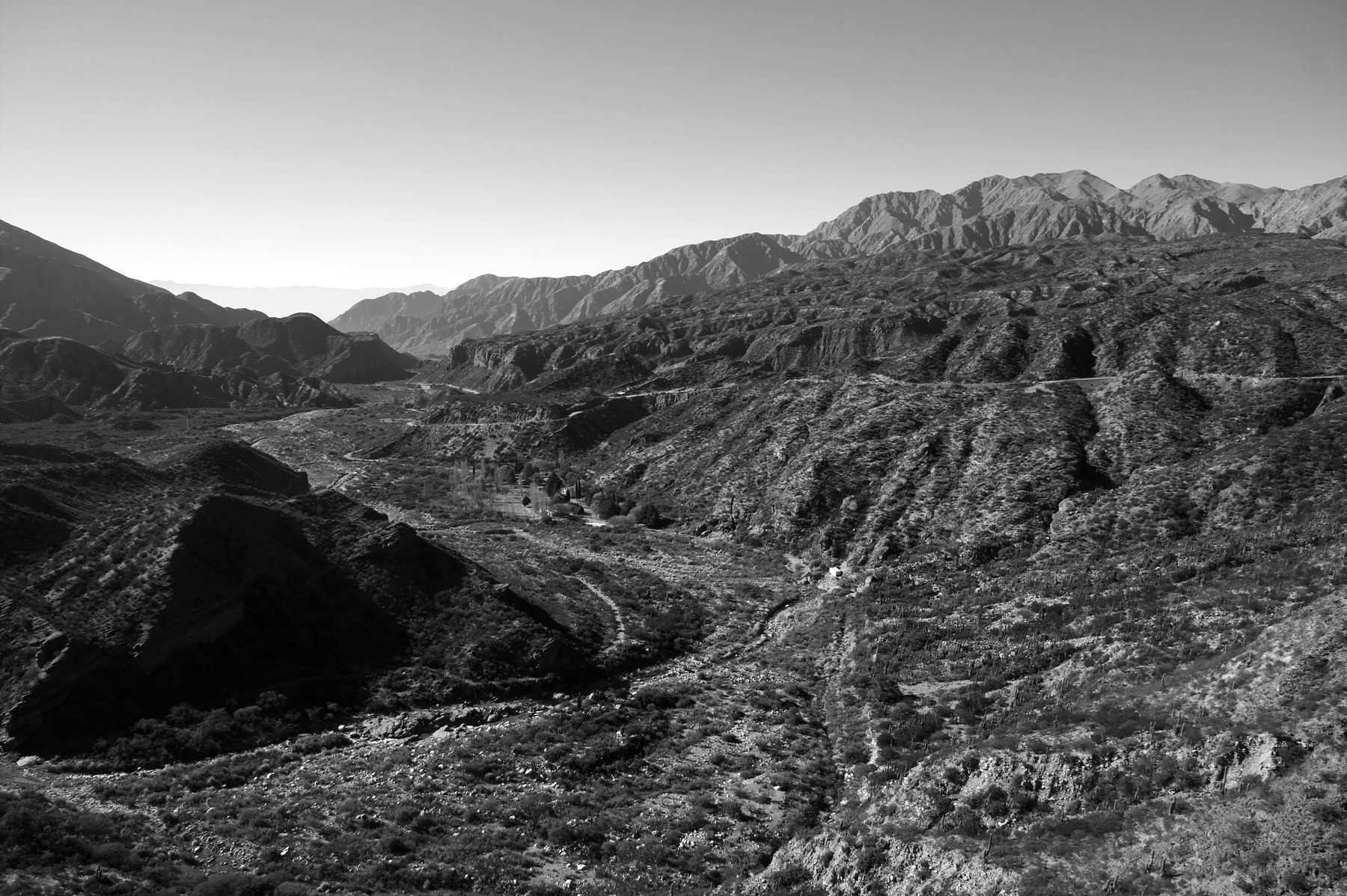 Looking down a long, dry valley of eroded hills and scrub, a thread of road and river bed winding through it towards distant ranges, under a clear sky.