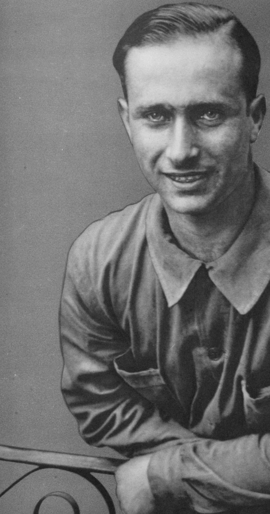 A half-length portrait of the young Fangio, smiling, his hair neatly combed, in a work shirt, his arms folded on an iron rail, against a plain background.