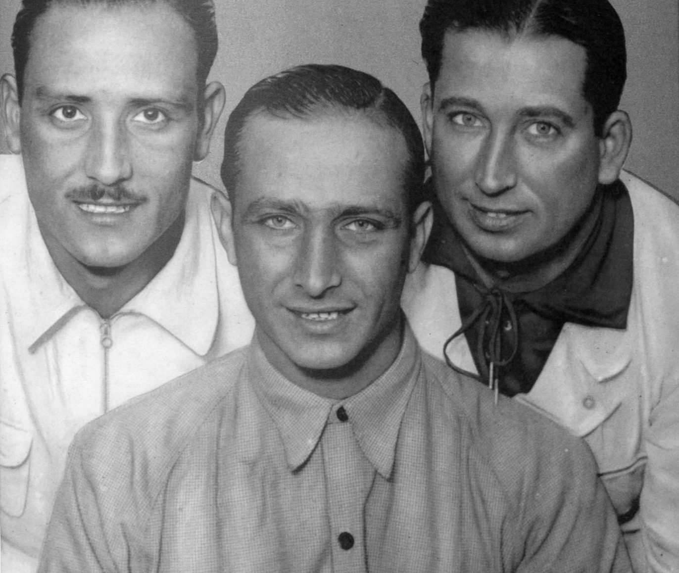 Three smiling men, head and shoulders, grouped close together: in front and centre Fangio in a work shirt; behind him on the left a man with a moustache in an open-necked shirt, on the right a man with a neckerchief knotted at his throat.
