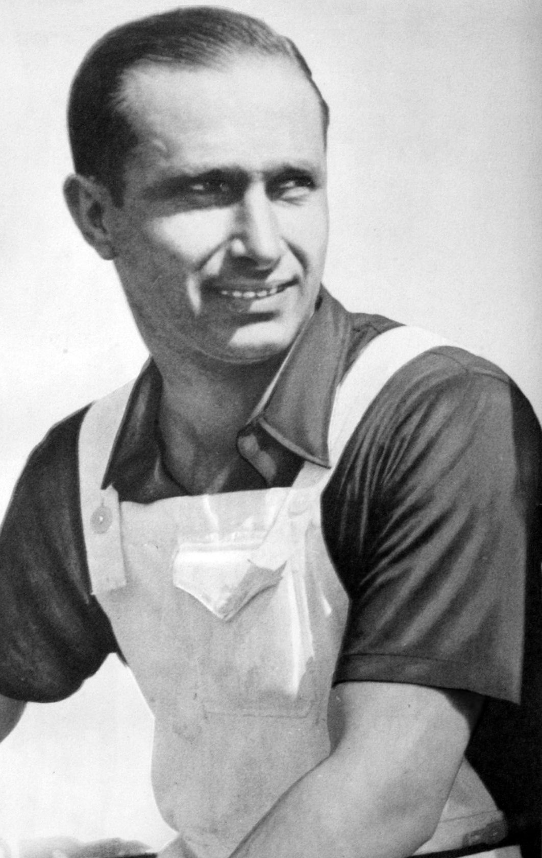 Fangio, smiling and glancing to one side, in a short-sleeved shirt under bib overalls, sits with his forearms resting across his knees.
