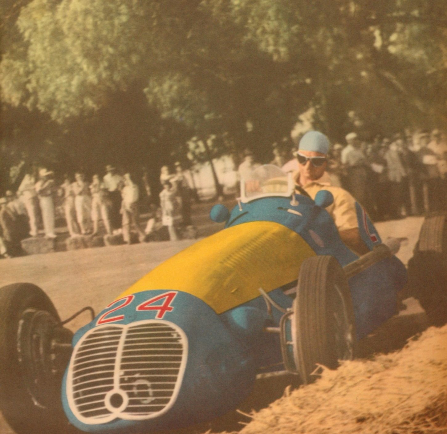 In colour: Fangio, in a pale helmet and goggles, drives a single-seater painted blue with a yellow bonnet and a red 24 on its nose past straw bales, spectators under the trees behind.
