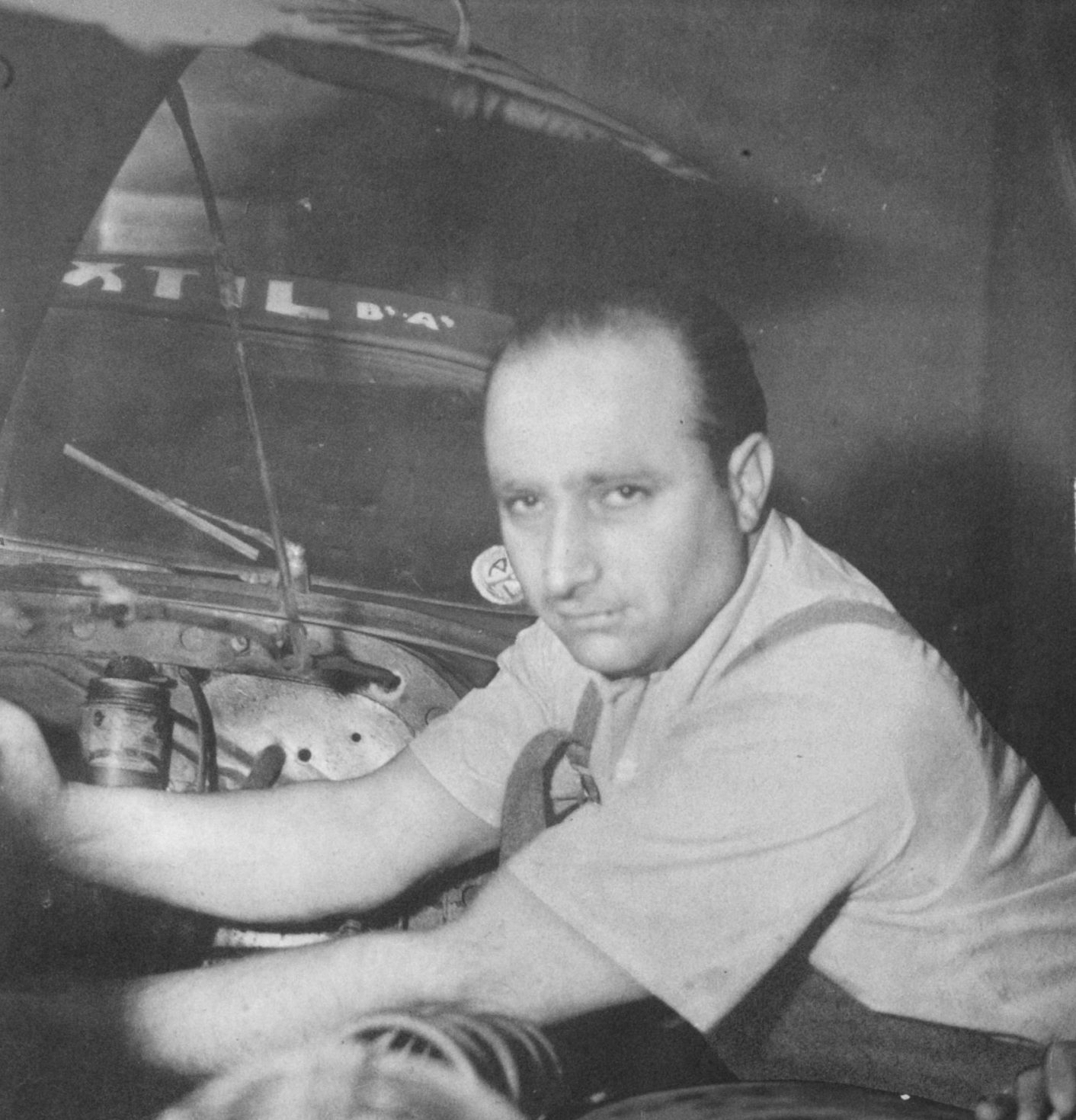 Fangio, in a pale short-sleeved shirt and braces, leans over the open engine bay of a car, its bonnet raised above him, and looks straight at the camera; the head of another man shows in the foreground.