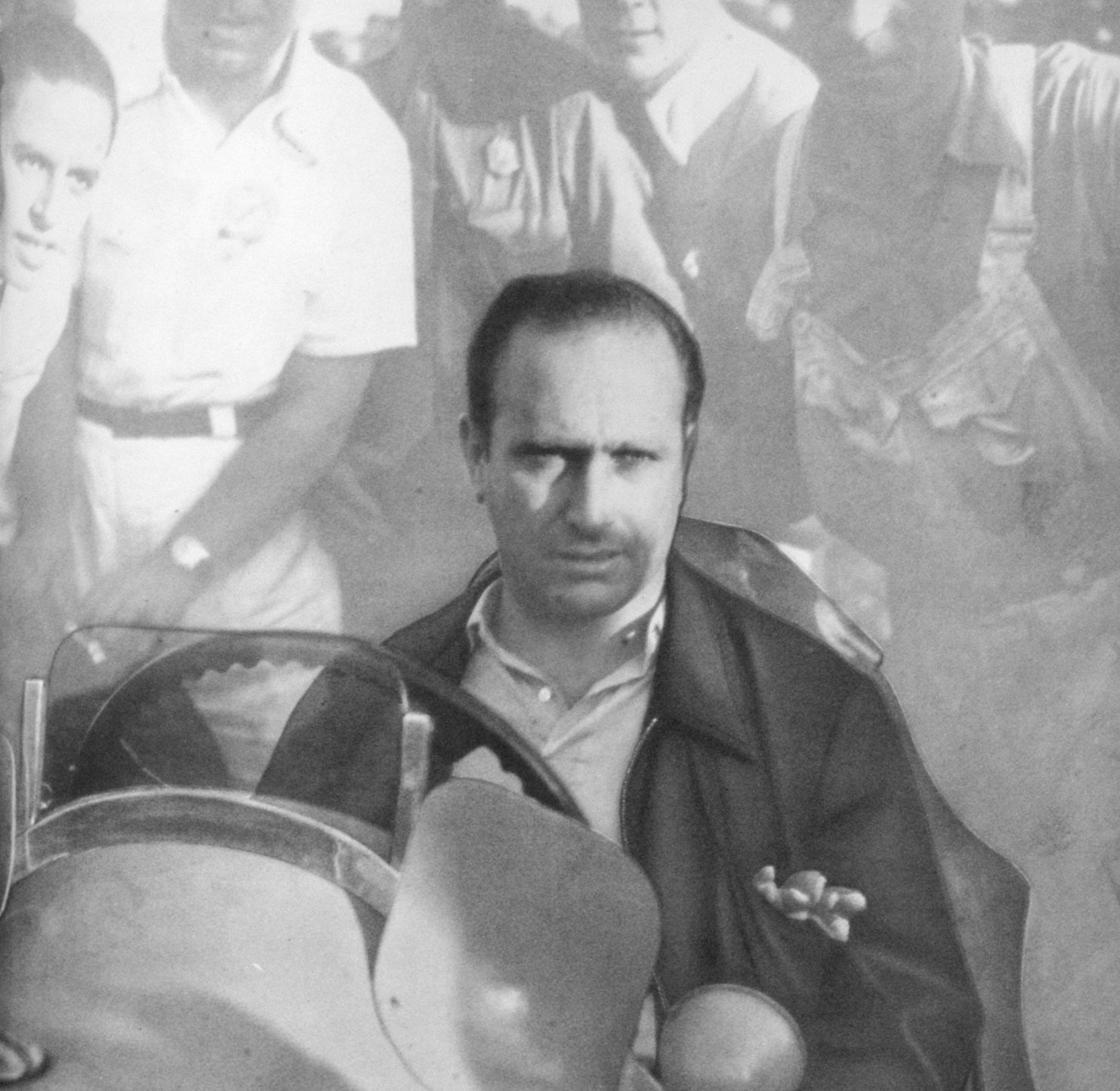 Fangio, unsmiling, in an open-necked shirt and a zipped jacket, sits behind the curved windscreen of a racing car; a crowd of men stands behind him.