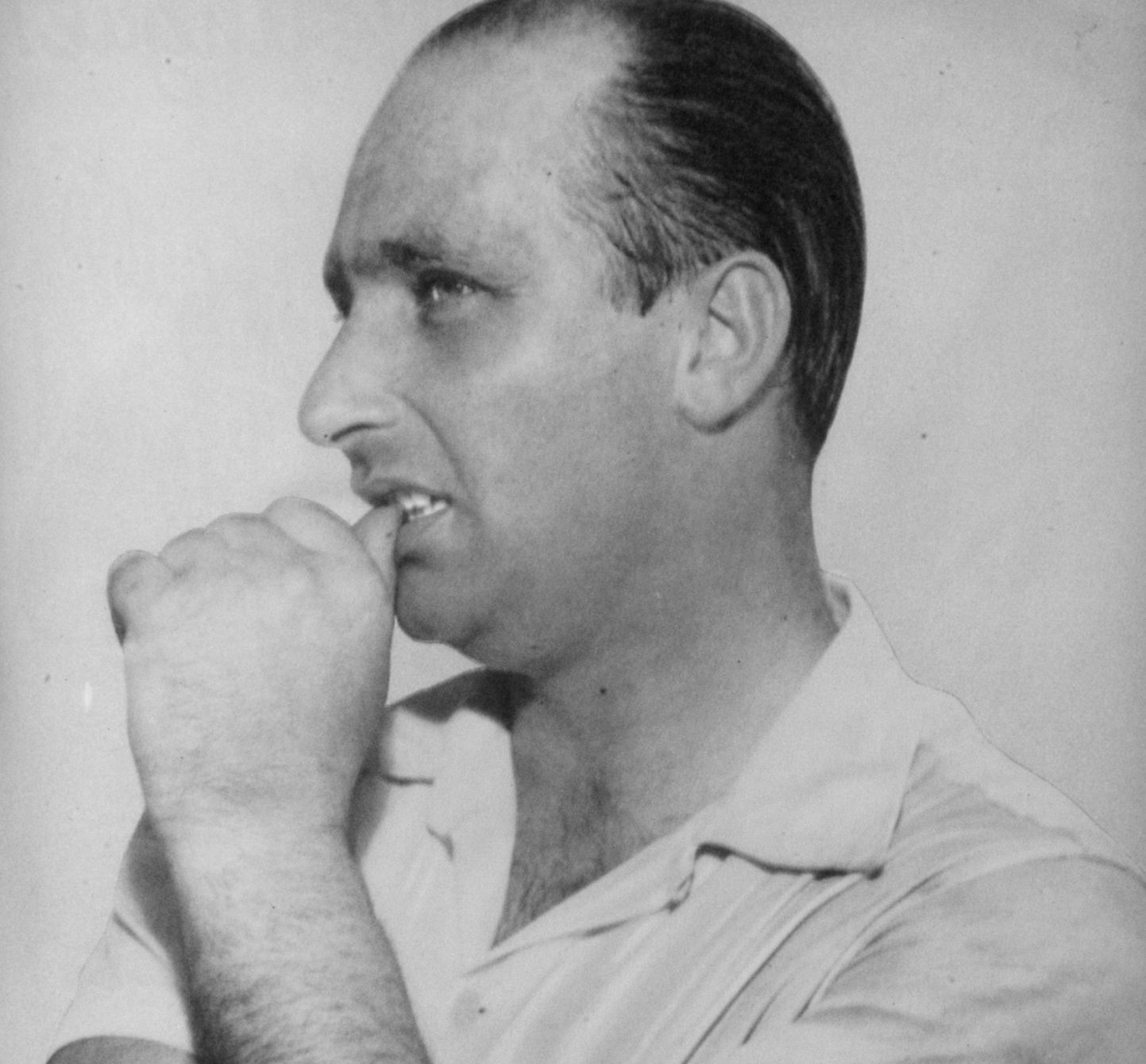 Fangio in profile, balding, his arms folded and a fist pressed to his lips as he looks ahead, in an open-necked short-sleeved shirt.