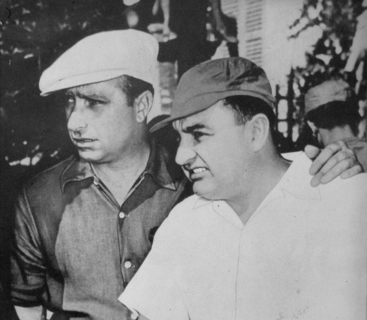 Two men side by side outdoors, both looking off to the left: Fangio in a pale cloth cap and a work shirt, and José Froilán González, heavier, in a darker cap and a white shirt, with a third man, partly hidden, resting a hand on González’s shoulder; foliage behind.