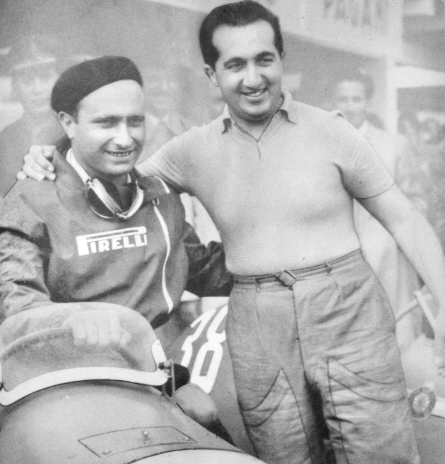 Fangio, in a beret and a zipped jacket lettered Pirelli, goggles round his neck, sits in a racing car numbered 38 and grins; Alberto Ascari stands beside the car in a short-sleeved shirt with an arm round his shoulders.