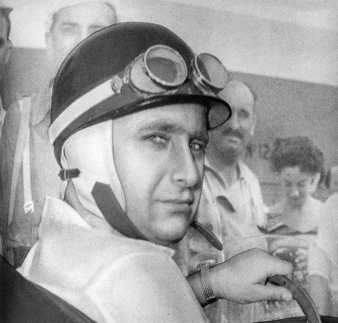 Fangio, seated at the wheel of a racing car, looks back over his shoulder at the camera; he wears a helmet with goggles pushed up on it, a cloth lining round his face and a loose chin strap, a short-sleeved shirt and a wristwatch. People stand behind him.