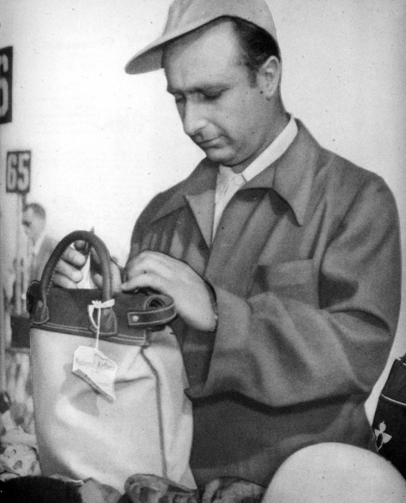Fangio, in a cloth cap and a zipped jacket, looks down as he packs a canvas kit bag with leather handles and a luggage label; a helmet lies in the corner and pit number boards reading 65 and 66 stand behind.