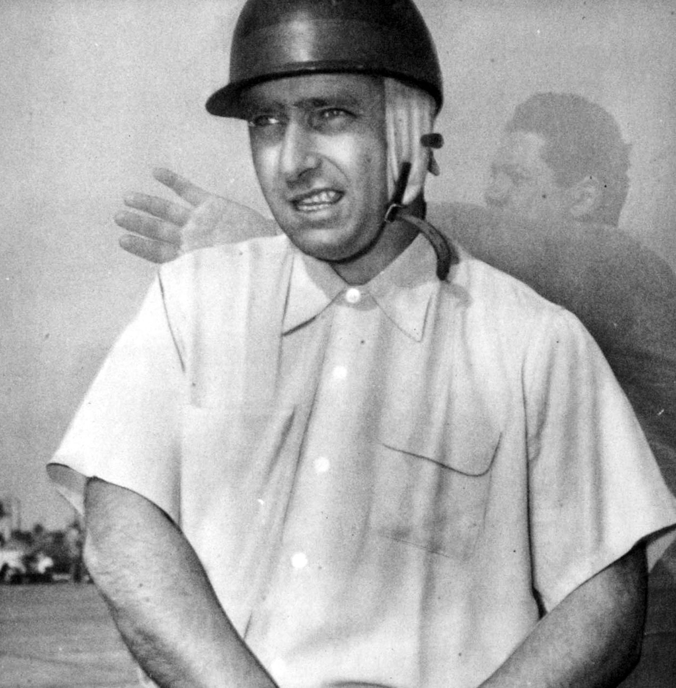Fangio, in a peaked hard helmet with a pale lining and a chin strap and a short-sleeved shirt, his arms low in front of him, looks to one side as he speaks; a blurred figure with a raised hand is behind him.