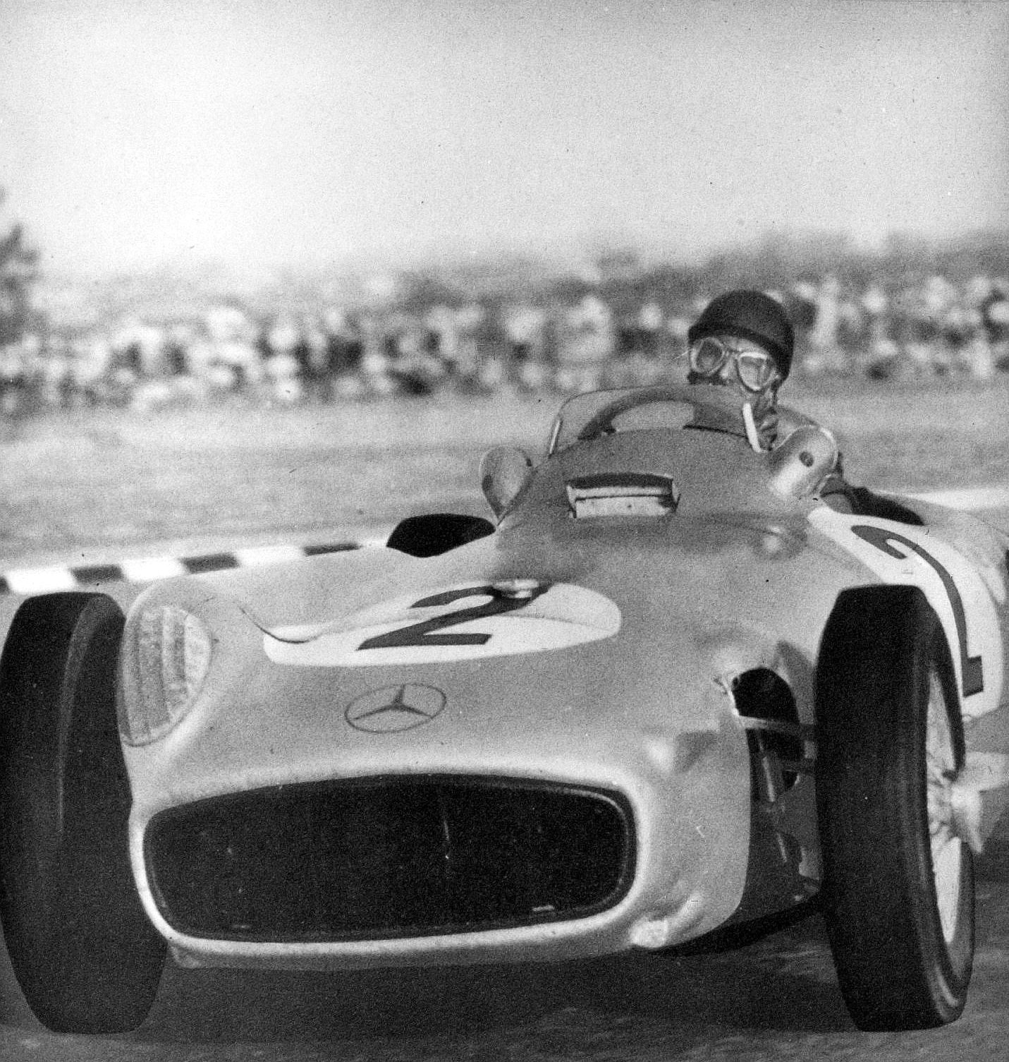 Seen head-on and low, Fangio in a helmet and goggles drives an open-wheeled Mercedes-Benz numbered 2 towards the camera, past a striped kerb, with grass and a blurred crowd behind.