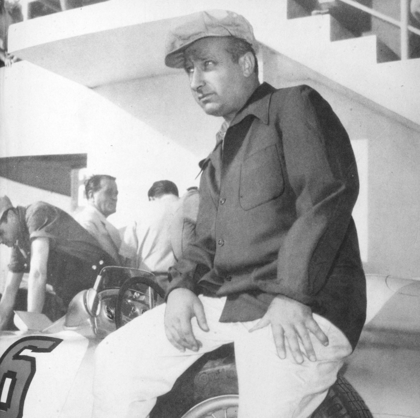 Fangio, in a cloth cap and a jacket buttoned over his shirt, sits on the front of a racing car numbered 6 with his hands on his knees and looks into the distance; men work at the car behind him, under the concrete steps of a grandstand.