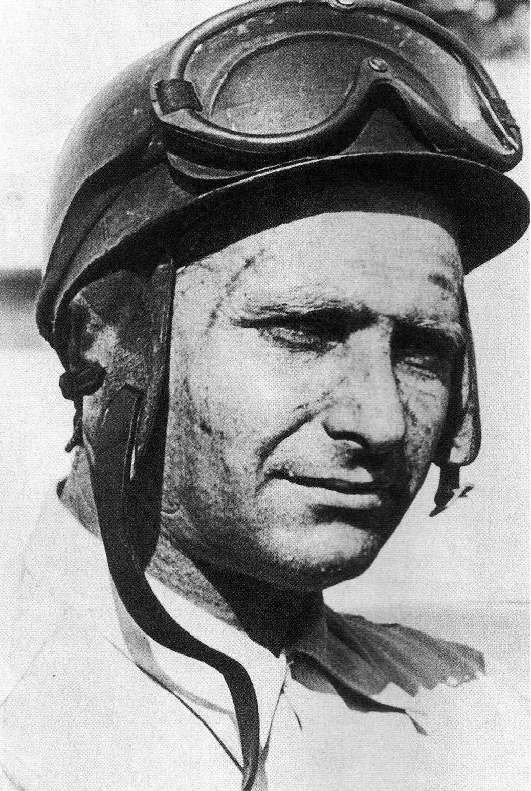 Fangio, head and shoulders, in a peaked hard helmet with ear flaps, the chin strap hanging loose and goggles pushed up on his brow; his face is streaked with road grime and he half-smiles, looking past the camera.