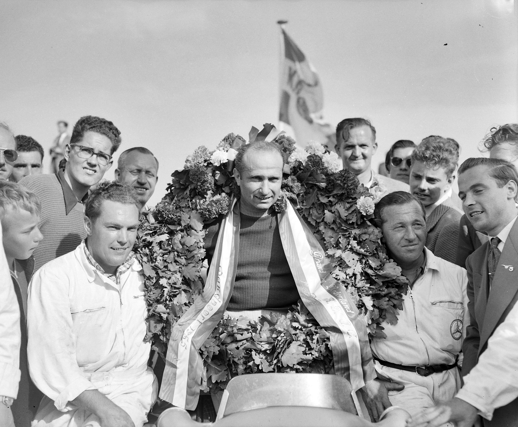 Fangio, behind a car’s windscreen, wears a huge garland of leaves and flowers with ribbons hanging from it, a flag flying behind; mechanics in pale overalls and grinning spectators crowd round him.