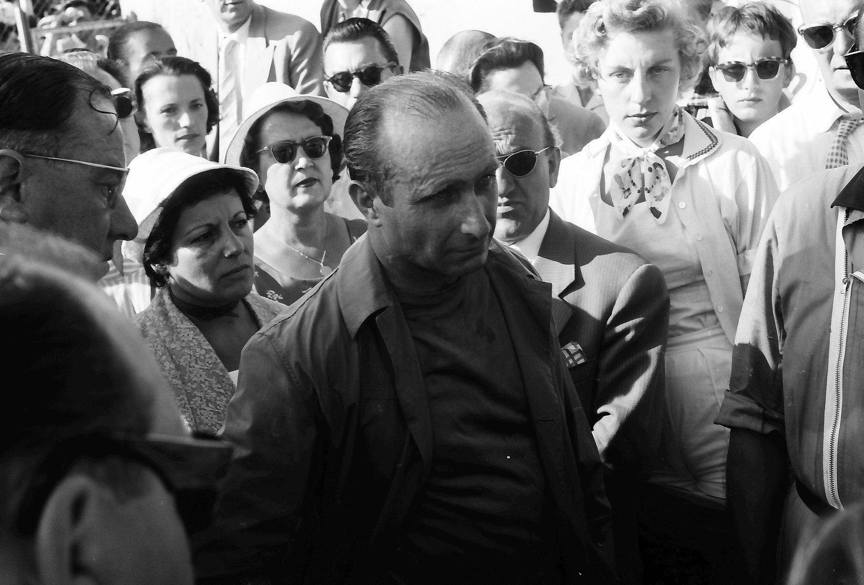 Fangio, drawn and unsmiling in an open jacket, moves through a crowd of spectators in sunglasses and summer hats, some of them turning to watch him.