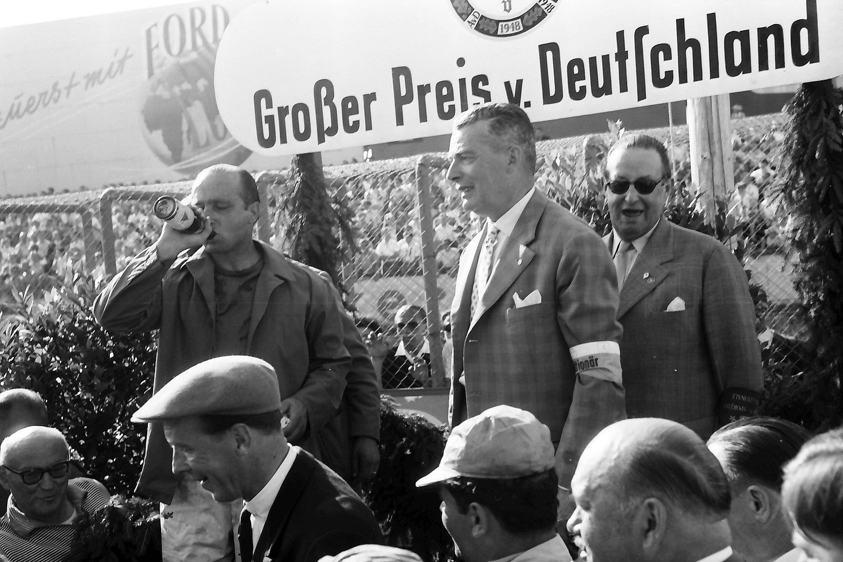 Fangio tips back a bottle under a banner reading Großer Preis v. Deutschland, beside officials in suits and sunglasses; a man in a flat cap laughs below.