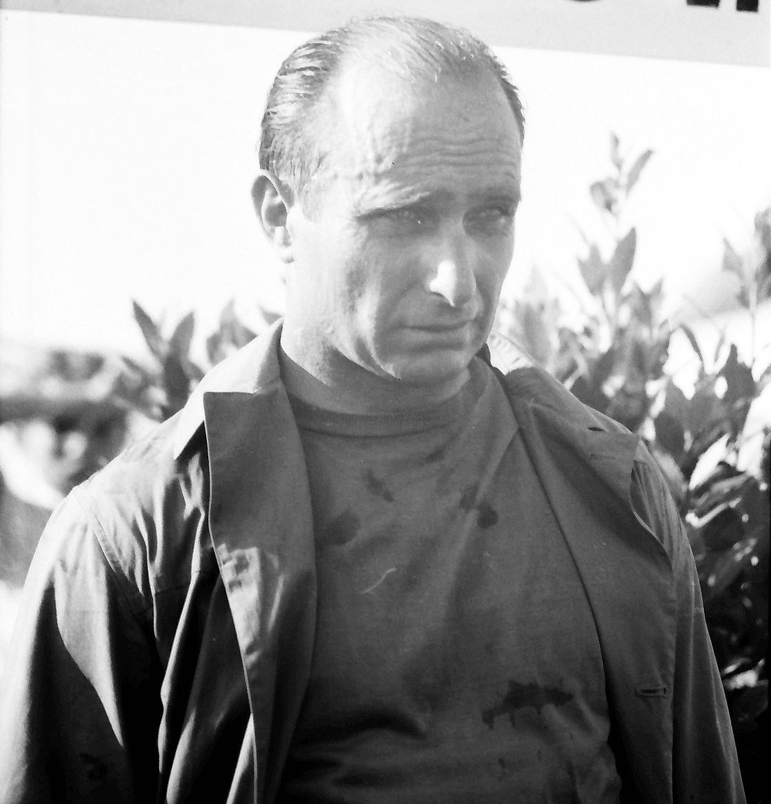 Fangio, eyes lowered, hair plastered down, stands drained and unsmiling in an open jacket over a stained dark shirt, leaves behind him.