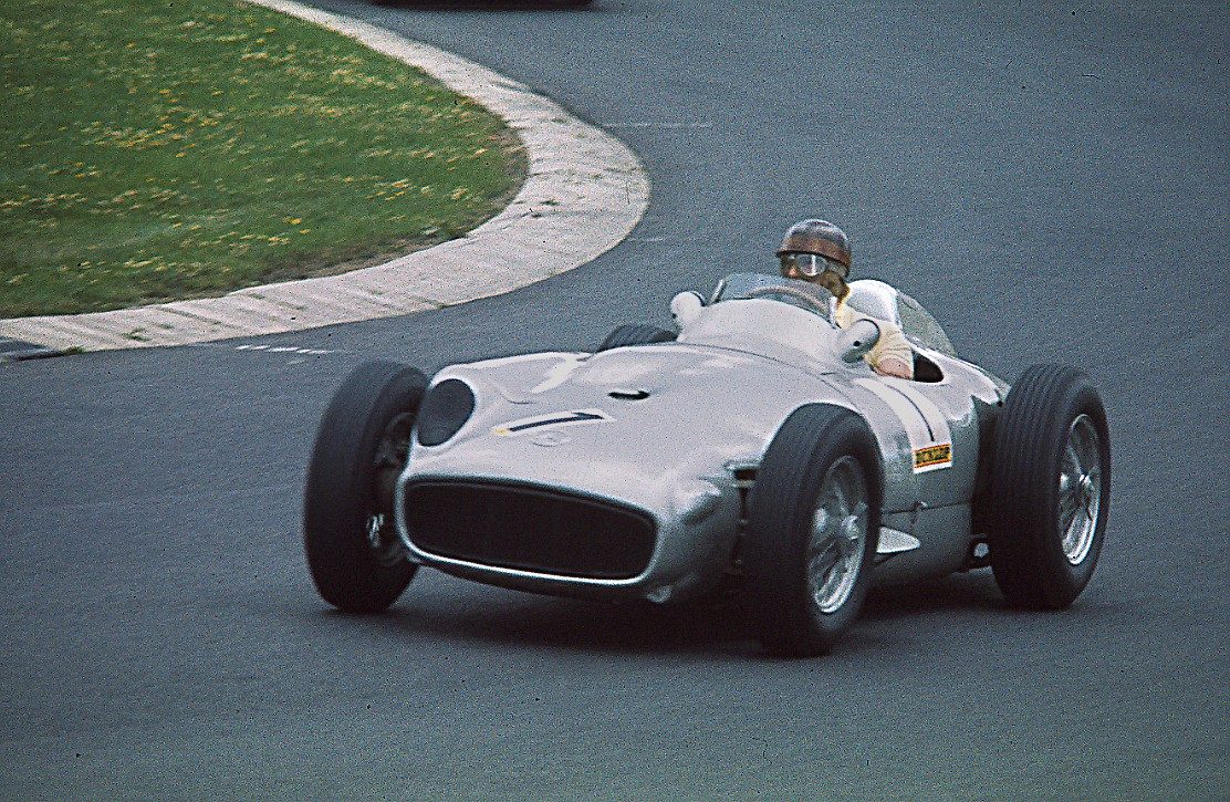 In colour: Fangio, in an open-face helmet and goggles, drives a silver Mercedes-Benz W196 open-wheeler through a kerbed bend, green grass inside the turn and grey tarmac beyond.
