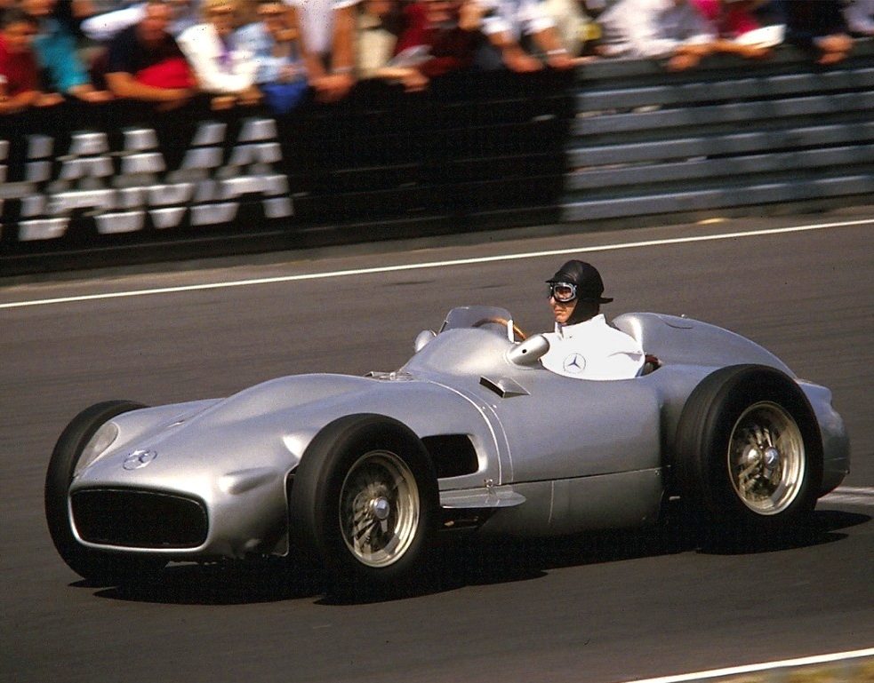 In colour: Fangio, in a dark open-face helmet and goggles, drives a silver Mercedes-Benz W196 along a straight, a crowd of spectators behind a barrier in the background.