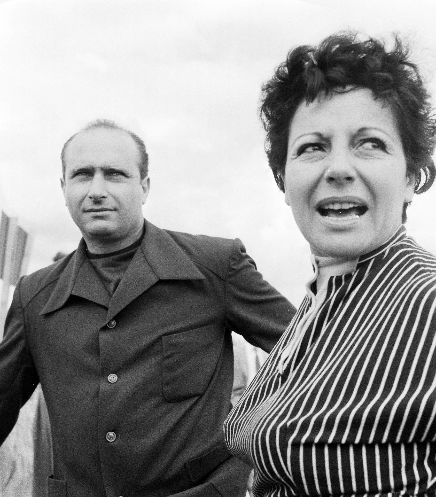 Fangio, bare-headed and balding, in a dark buttoned jacket over a roll-neck, looks off to the left with a level stare; beside him a woman with short curly hair and a striped blouse turns to speak, mouth open. Overcast sky behind.