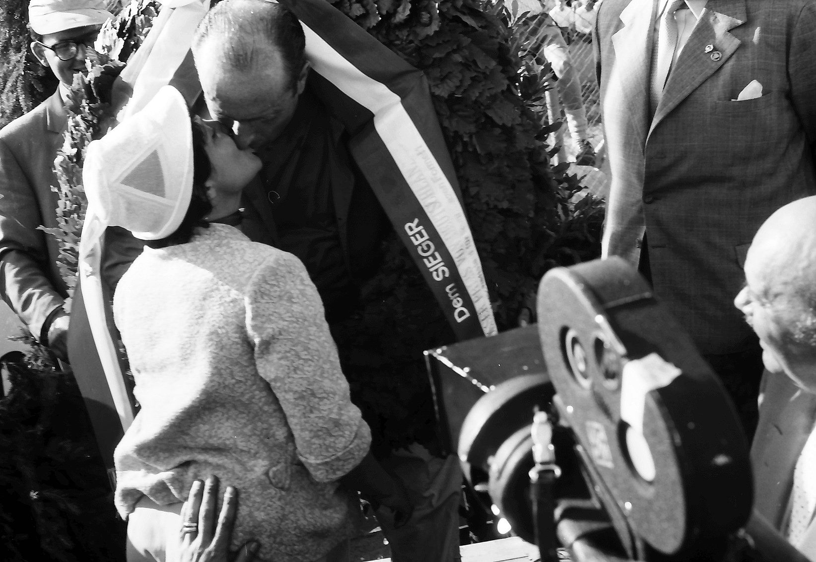 On the podium Fangio, a sash lettered Dem Sieger across his chest, bends to kiss a woman in a pale headscarf and knitted jacket; a newsreel camera looms in the right foreground and officials stand close.