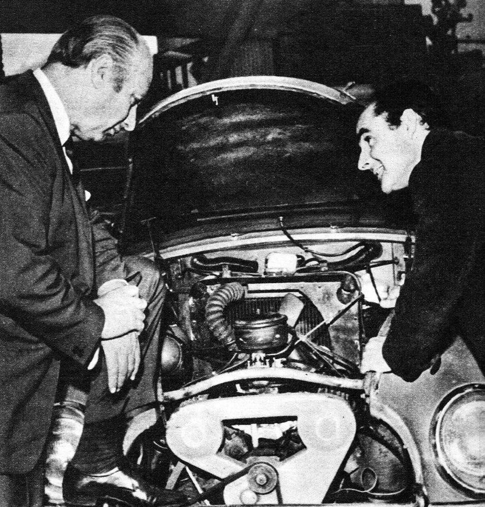 A magazine halftone: a grey-haired man in a suit, on the left, leans in over the open bonnet of a small car to look at its engine, while a young man opposite explains it, smiling.