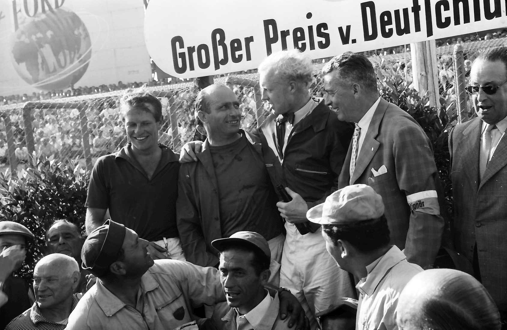 Three drivers laugh together under a banner reading Großer Preis v. Deutschland: a young man in a dark shirt on the left, the balding Fangio in the centre and a fair-haired man turning to an official on the right; mechanics and photographers crowd below.