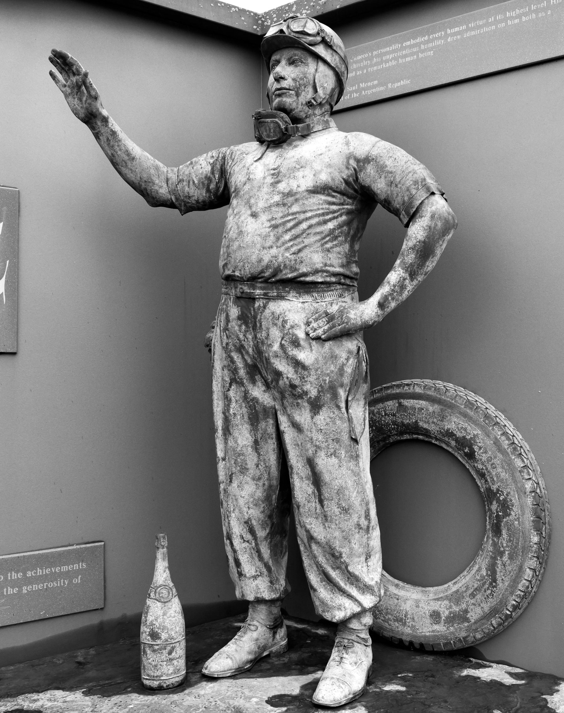 The bronze Fangio of the Donington memorial: goggles pushed up on a cloth helmet, right arm raised in greeting, left hand on his hip, a short-sleeved shirt and baggy trousers, a tyre and a bottle beside him; a weathered patina.