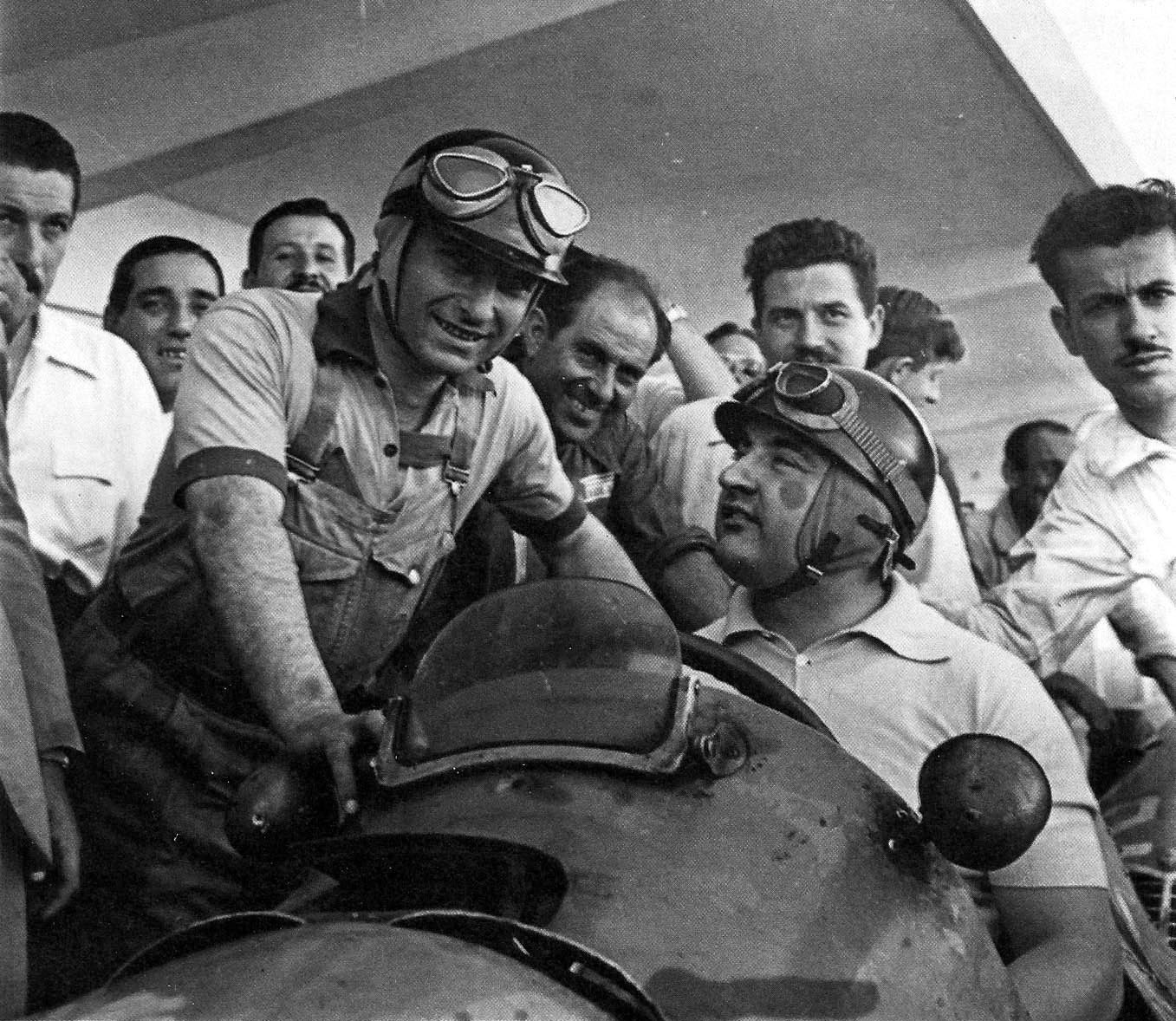 Under a concrete roof, Fangio, in helmet, goggles and a short-sleeved work shirt with braces, leans grinning over the cockpit of a racing car, where José Froilán González sits in helmet and goggles looking up at him. Mechanics and onlookers press round them.