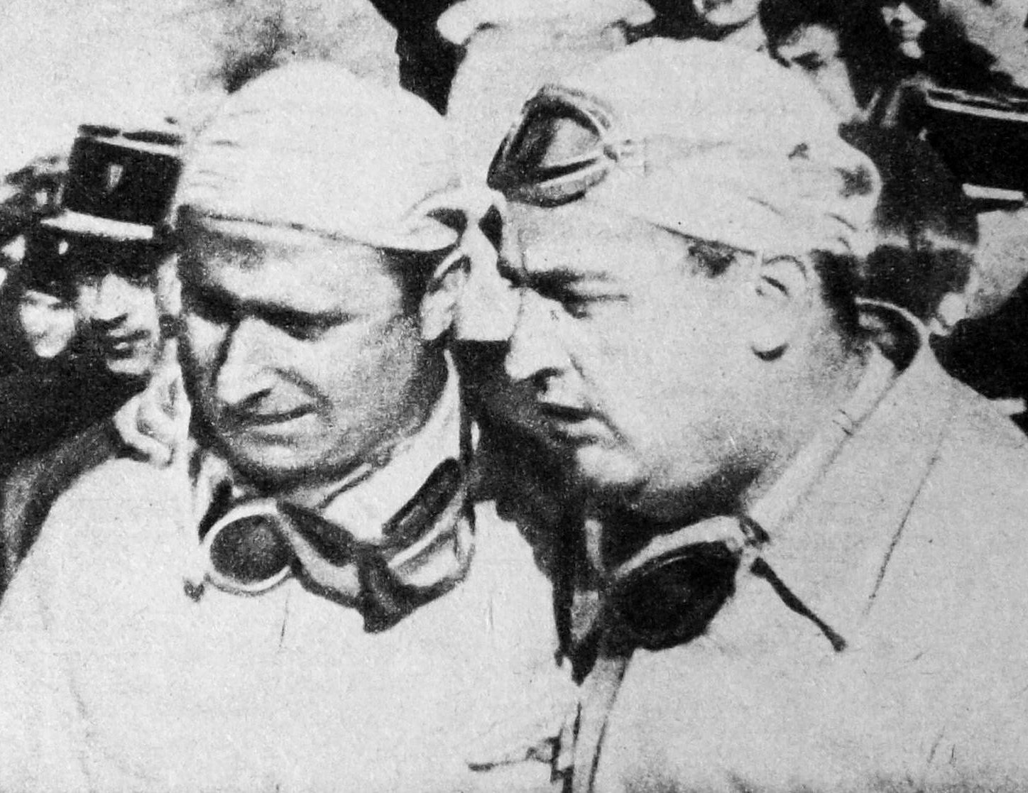A coarse newspaper halftone: two men in pale cloth caps, goggles round their necks, bow their heads together as they talk, a policeman in a peaked kepi behind them in a crowd.