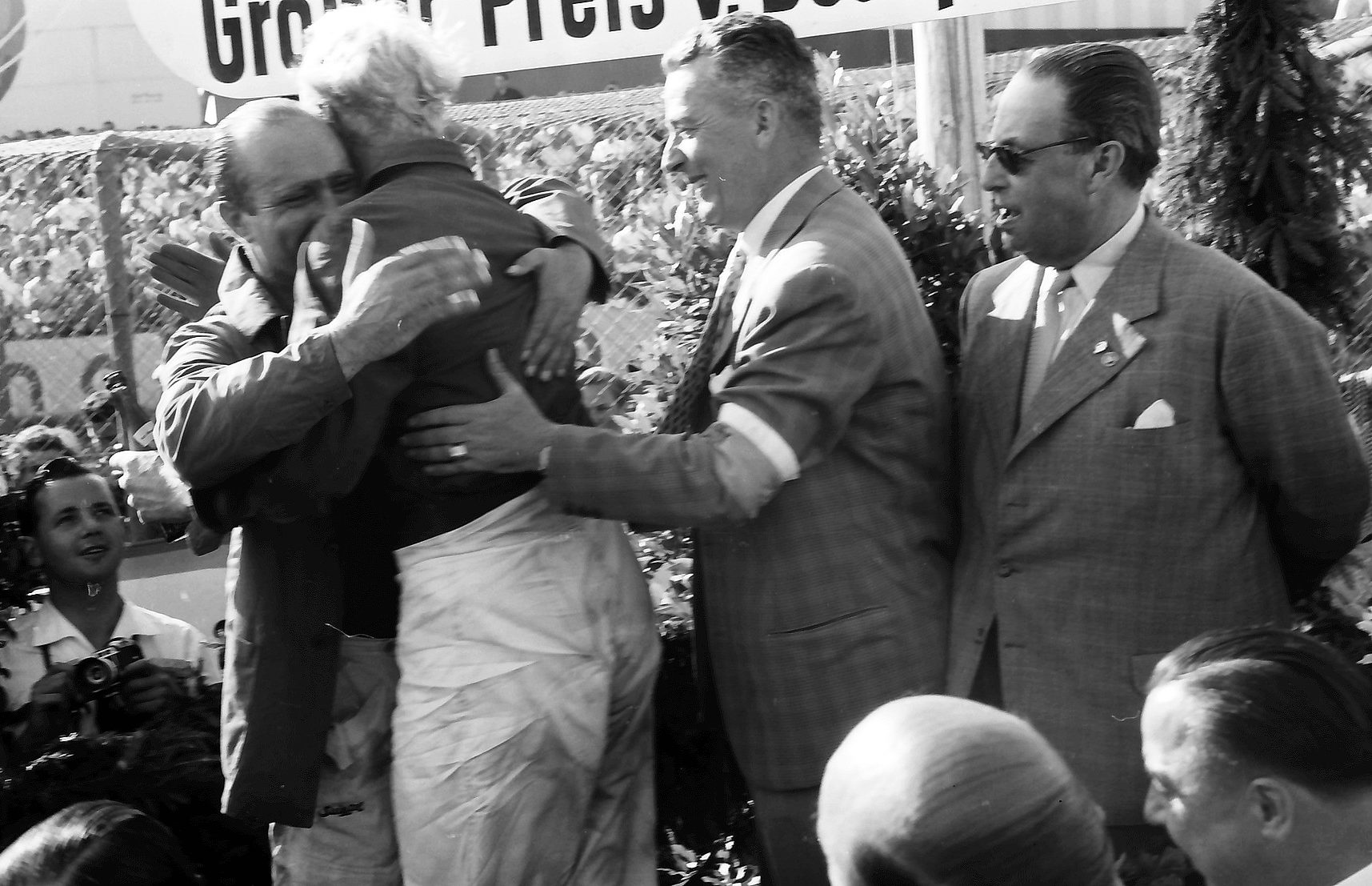 On the podium a fair-haired driver, his back to the camera, throws his arms round Fangio, whose eyes are closed; an official with an armband and a man in sunglasses look on, a press photographer at the lower left.
