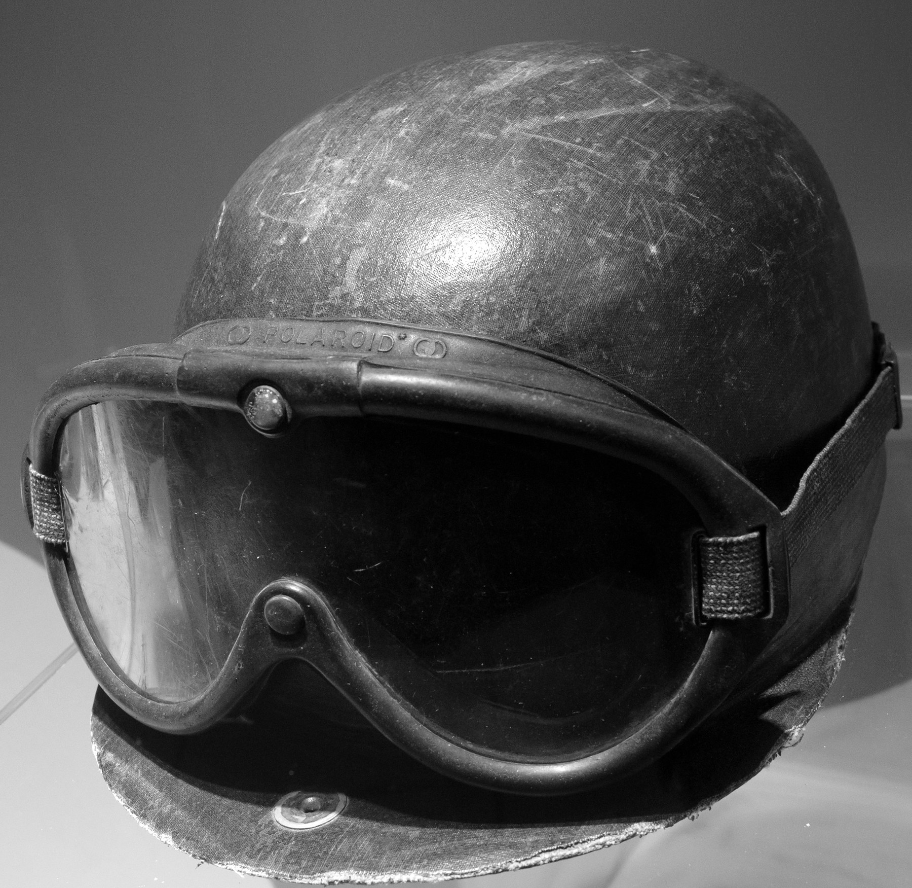 A worn, peaked hard helmet covered in scuffed and scratched leather, with rubber-framed goggles stretched round it on a fabric strap, in a museum case.