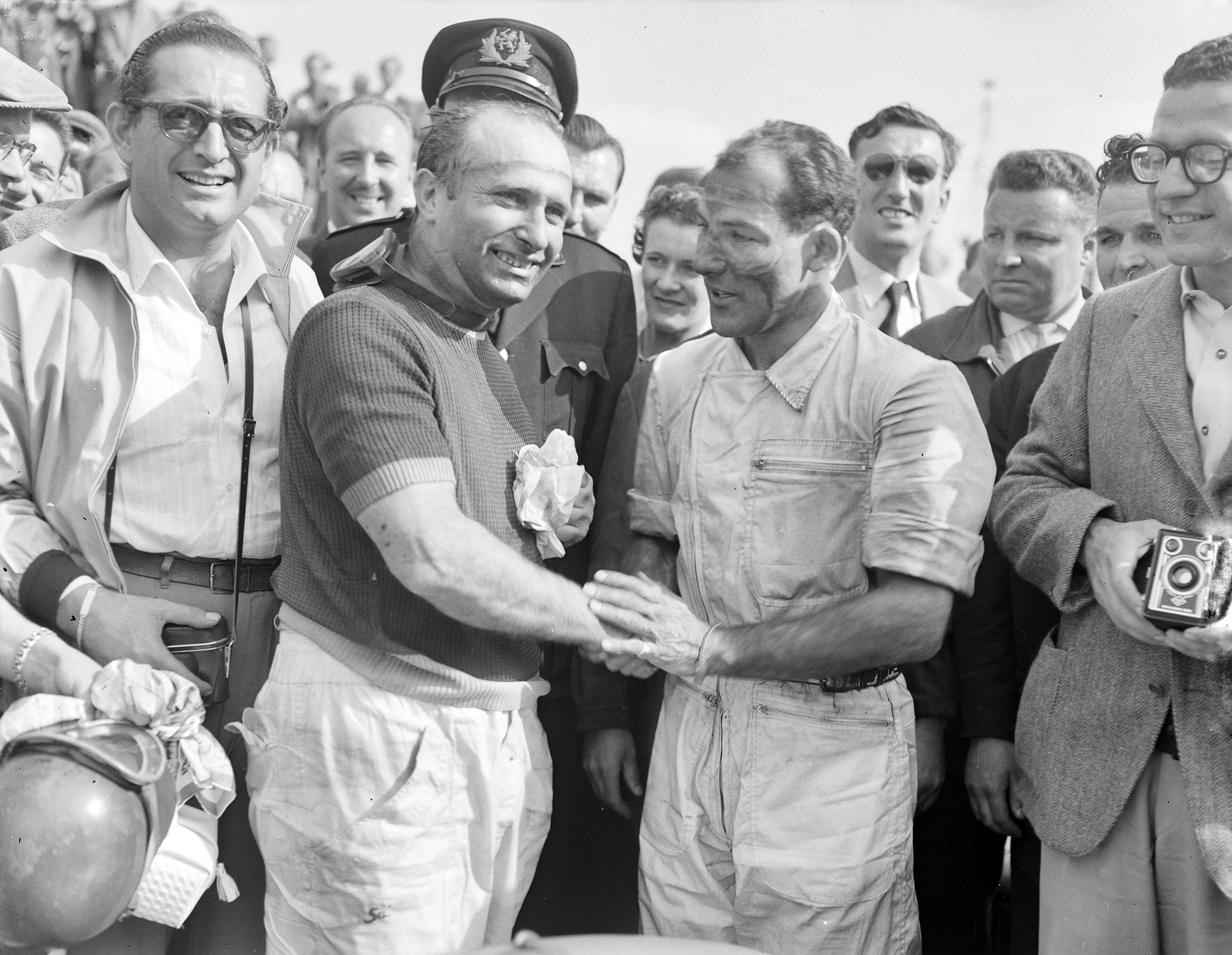 Fangio, in a knitted short-sleeved shirt and smiling broadly, shakes hands with Stirling Moss, grimy-faced in overalls; officials, a policeman in a peaked cap, a man in spectacles and another holding a box camera crowd round them.