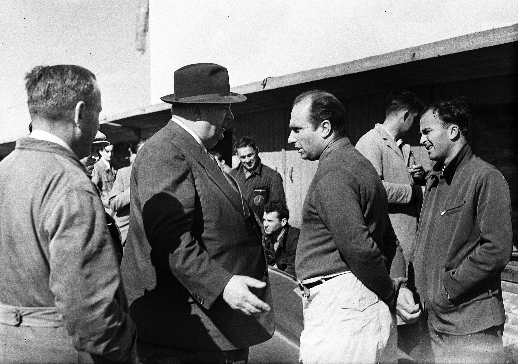 In the pit lane a stout man in a trilby and overcoat, back half turned, gestures as he talks to Fangio, who stands in a dark knitted shirt and pale trousers; a mechanic in overalls at the left, a man in a zipped jacket grinning at the right, the pit roof behind.