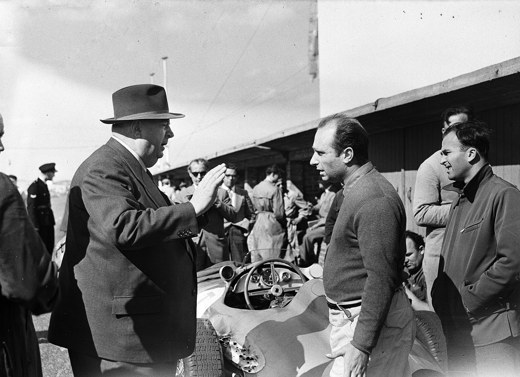 A second frame: the man in the trilby raises an open hand as he makes a point; Fangio listens with his arms at his sides, beside the cockpit of a racing car whose steering wheel and dials show in the foreground; a policeman and onlookers behind.