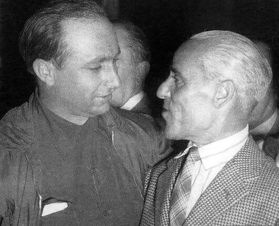 Two men face to face against a dark background: on the left Fangio, in a dark shirt, smiling down; on the right the older Tazio Nuvolari in profile, grey-haired and lean, in a checked jacket and striped tie.