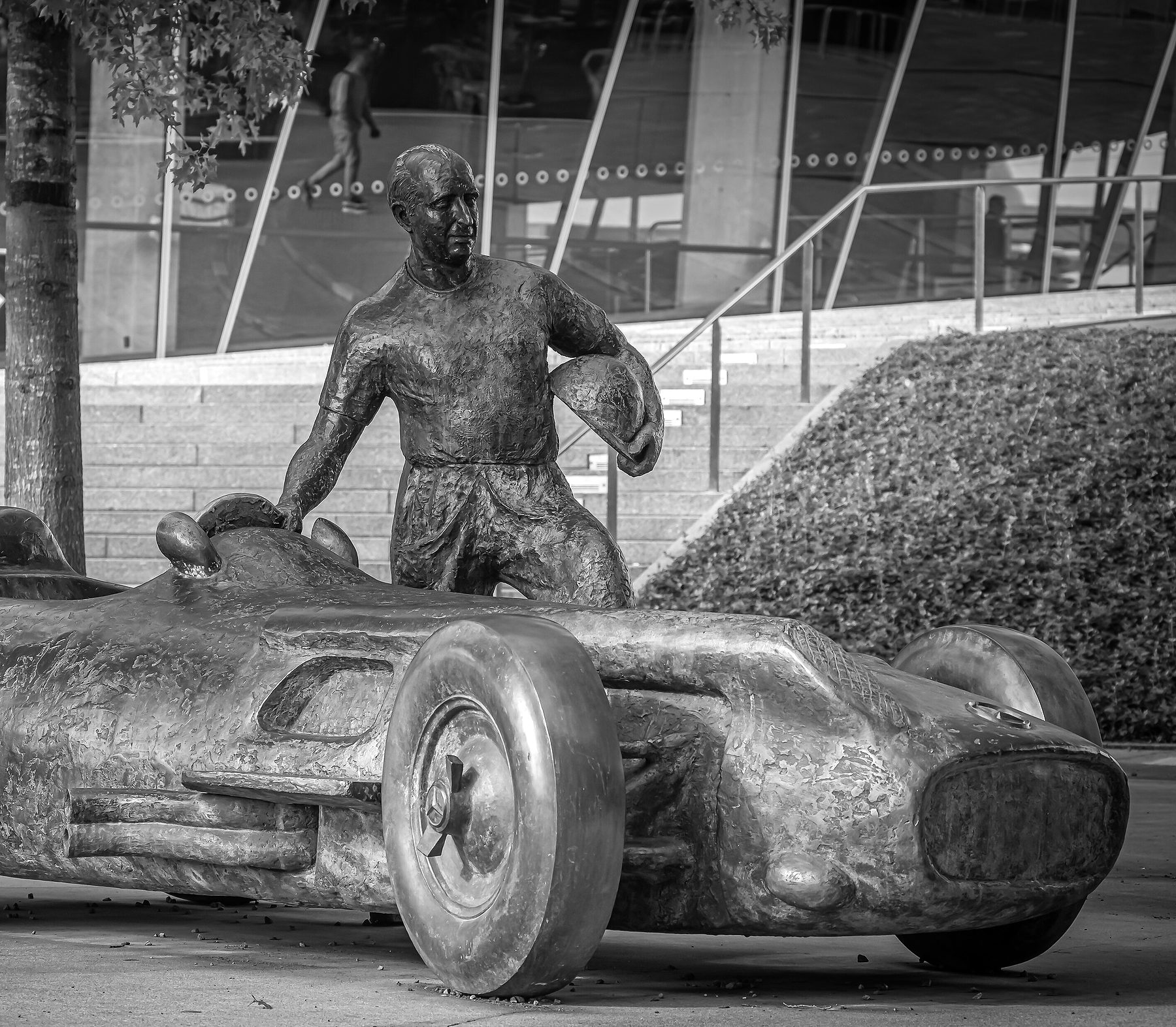 A life-size bronze of Fangio standing beside an open-wheeled racing car, his right hand on its steering wheel and a helmet under his left arm; behind him the steps and glass front of a museum, a hedge bank, and one passer-by walking along the terrace.