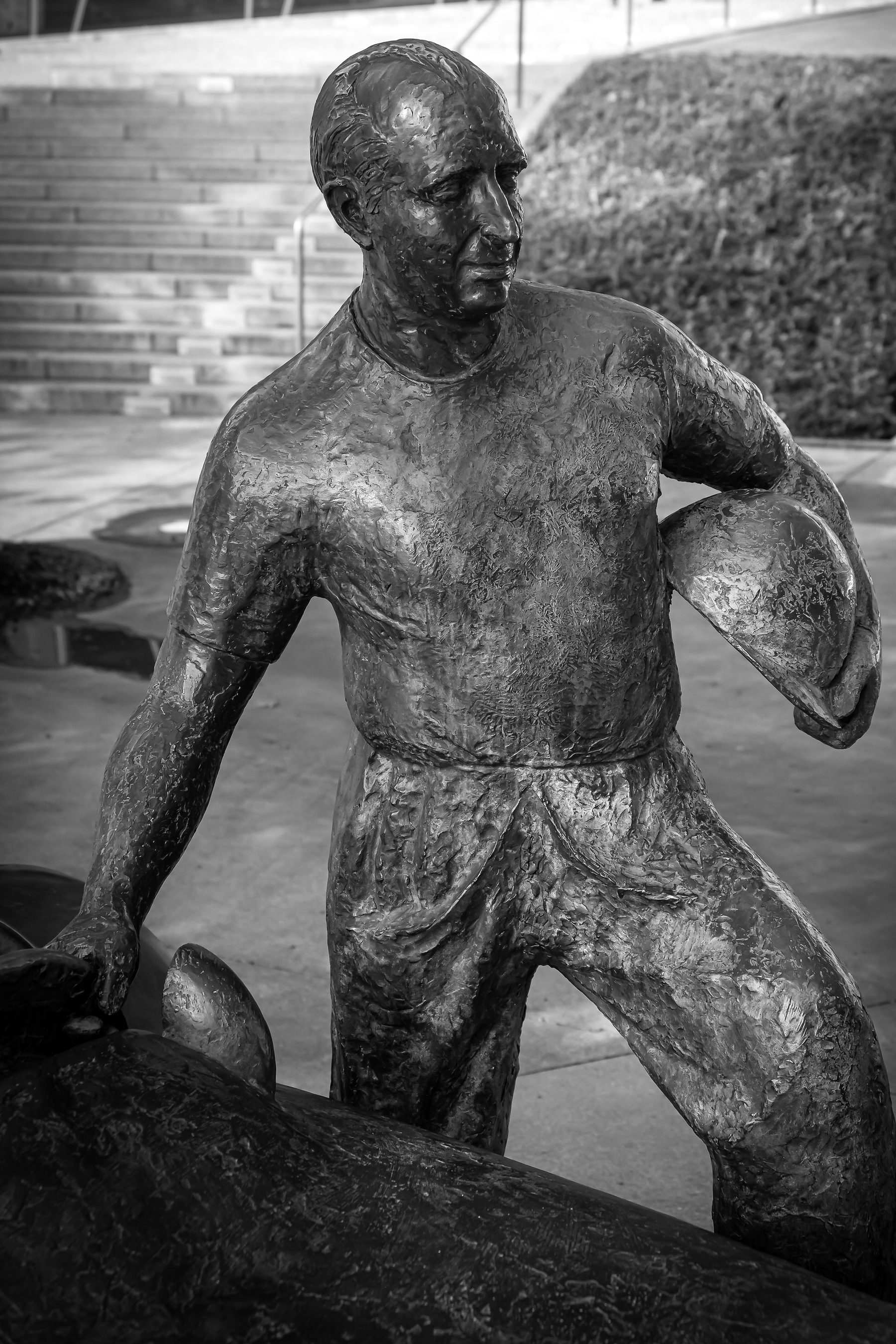 The bronze figure of Fangio, close, from the knees up: a short-sleeved shirt and creased trousers, his head turned to one side, a helmet held against his hip in one hand and the other hand resting on the car; museum steps and a hedge bank behind.