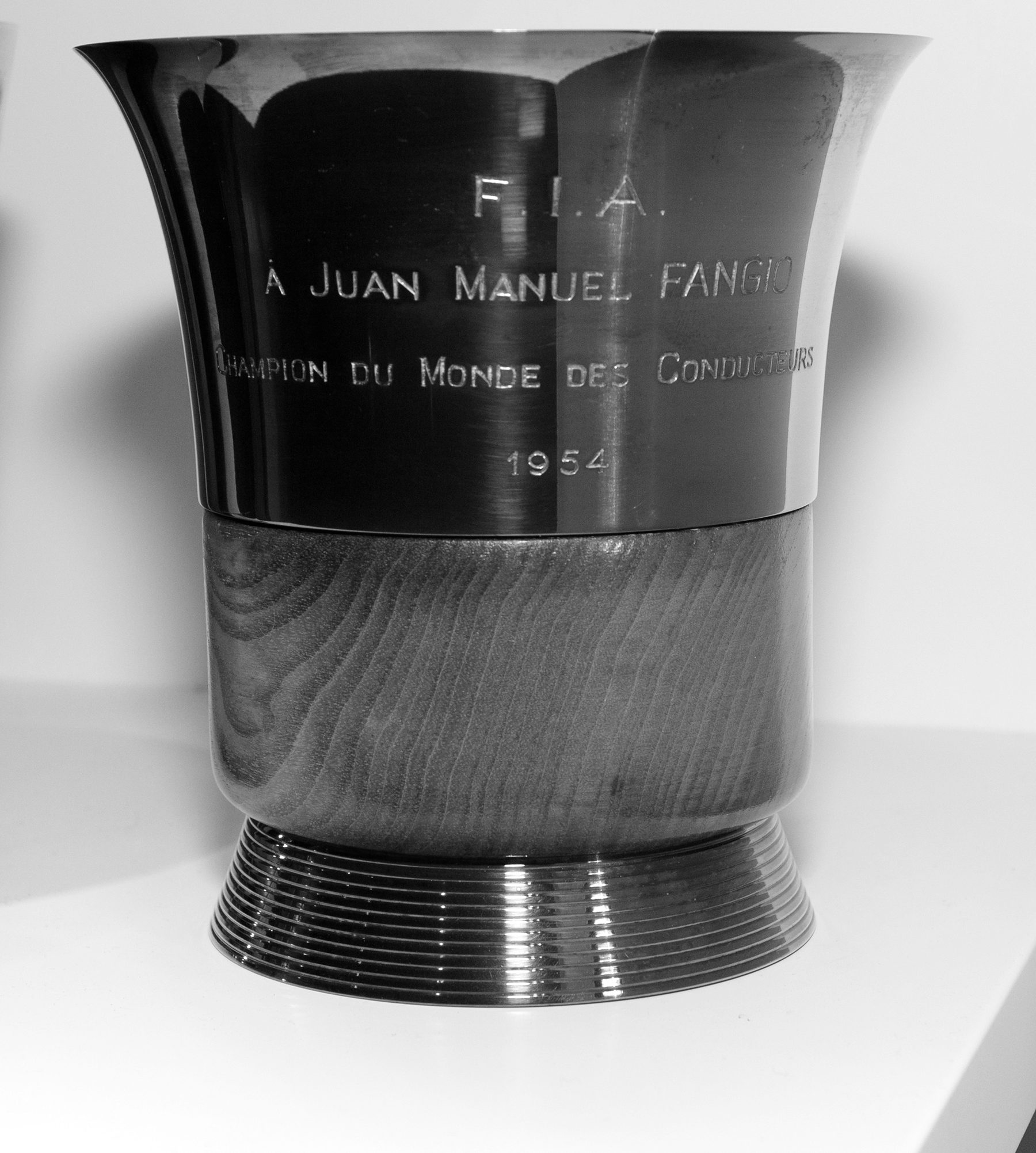 A plain beaker-shaped metal cup on a turned wooden base and a ribbed foot, engraved F.I.A., À Juan Manuel Fangio, Champion du Monde des Conducteurs, 1954.
