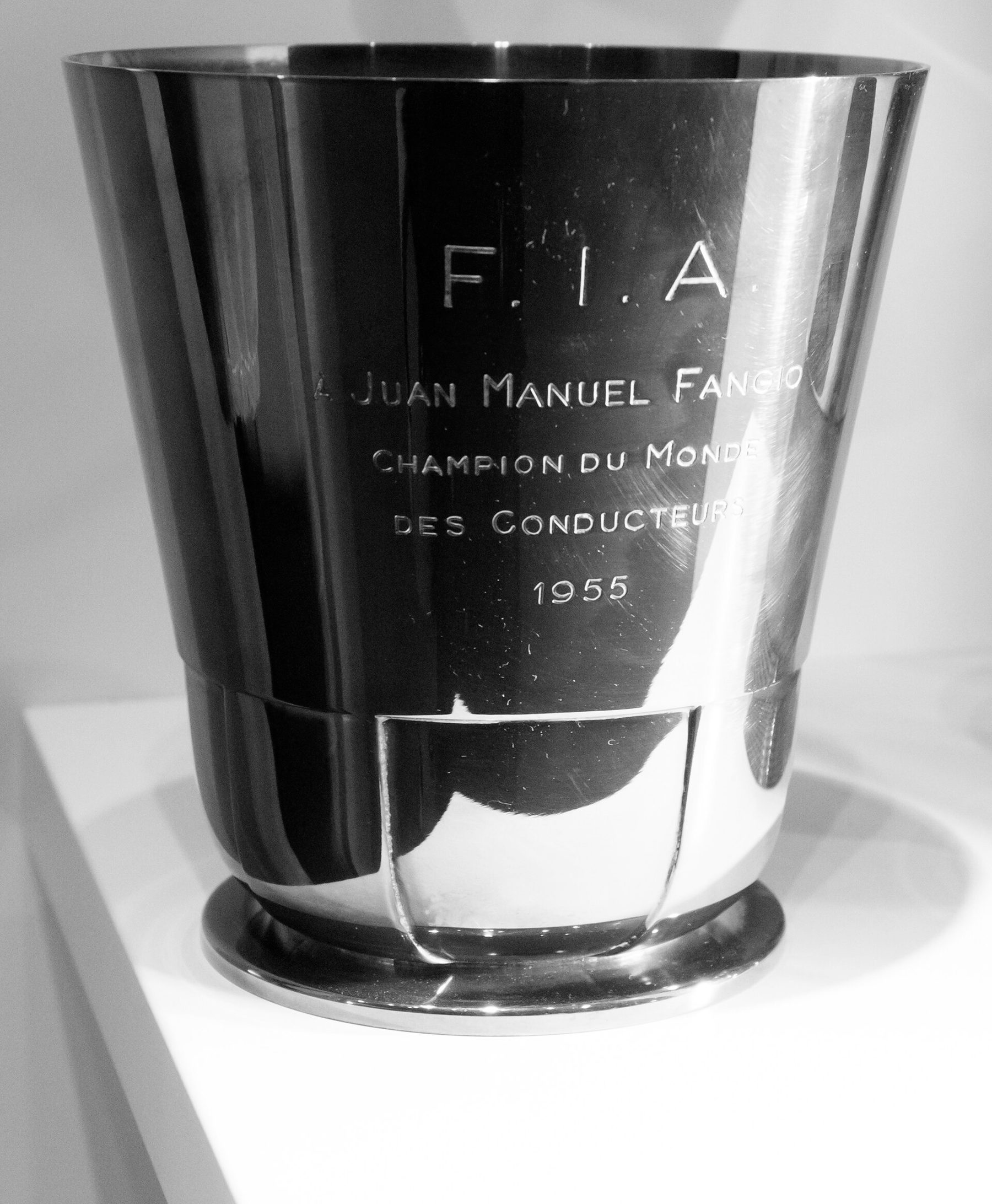 A polished, flared metal beaker, strongly reflective, engraved with the FIA's initials, Fangio's name and the year 1955, on a white plinth.